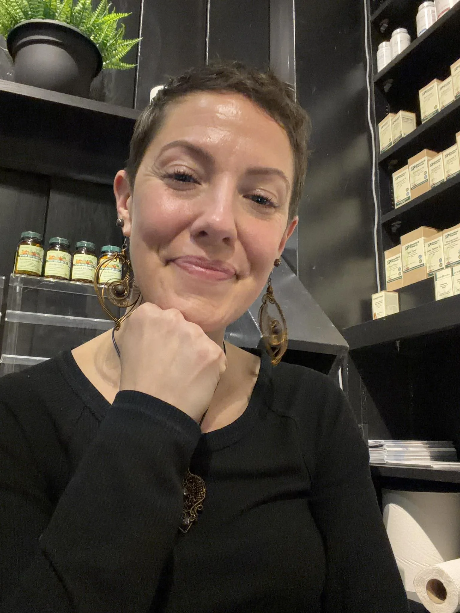 A woman with short, dark hair smiling and resting her chin on her hand, sitting in a room with black shelves filled with health products, a potted plant on top, and a roll of toilet paper nearby.