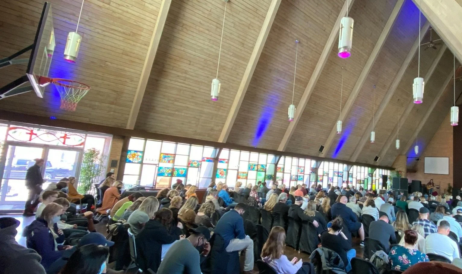 A large group of people seated inside a church during a service, with a stage at the front and stained glass windows along the side wall. The ceiling is high and wooden, with hanging pendant lights.