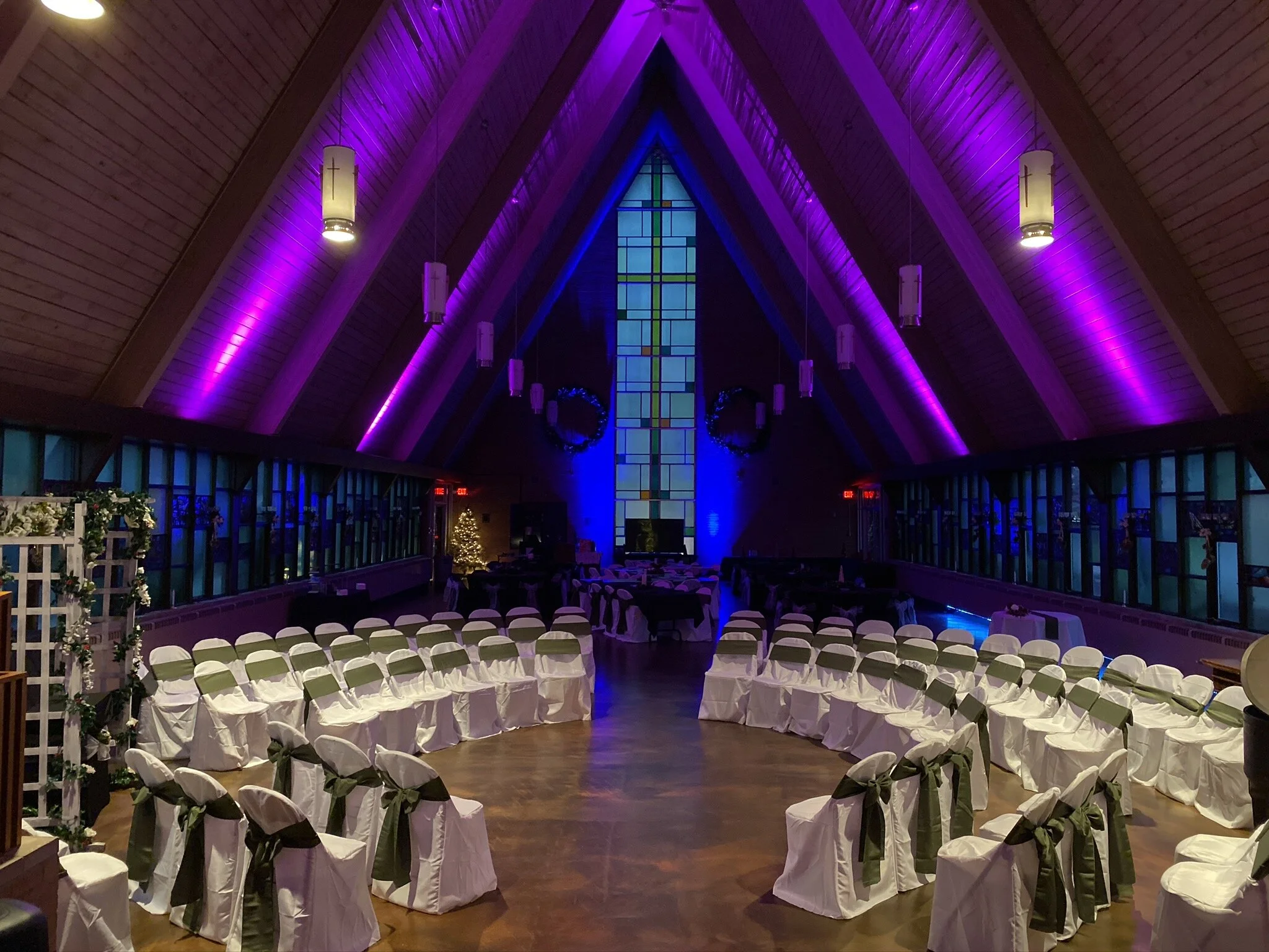 Interior of a church decorated for a wedding with rows of white chairs with black sashes, a large stained glass window with a cross, purple and blue lighting, and wreath decorations.