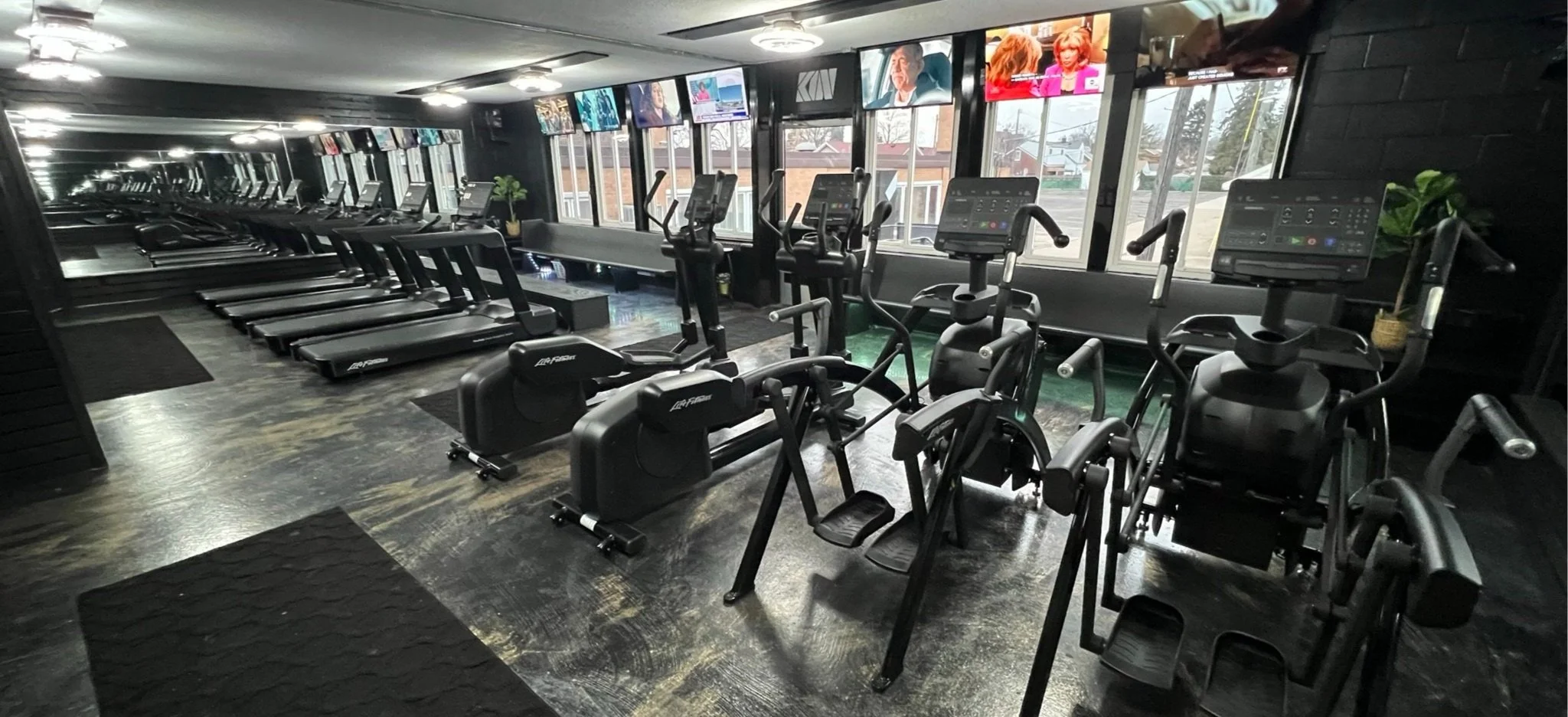 A modern gym with treadmills, ellipticals, and stationary bikes, with large windows and multiple TV screens mounted on the wall.