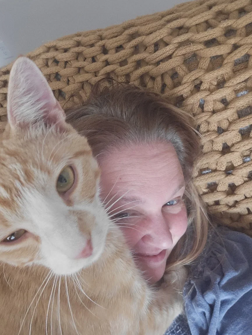 A woman with light brown hair lying down on a woven surface, smiling, with an orange and white cat close to her face, both looking at the camera.