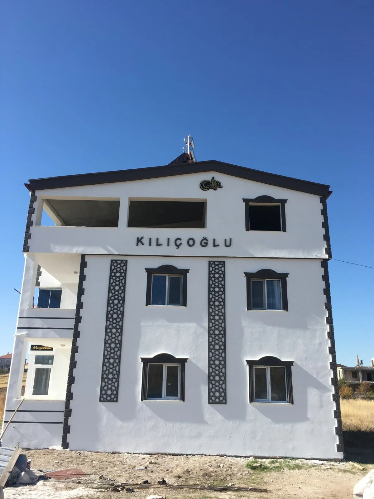 A white, multi-story building with black decorative accents and the word 'KILIÇOĞLU' on the front. It has several windows and an open balcony on the top floor, with a clear blue sky in the background.