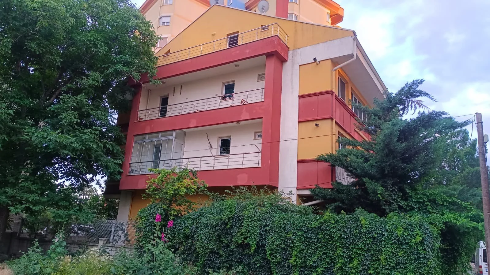 A multistory apartment building painted in yellow, orange, and white with balconies and railings, surrounded by green trees and shrubs.