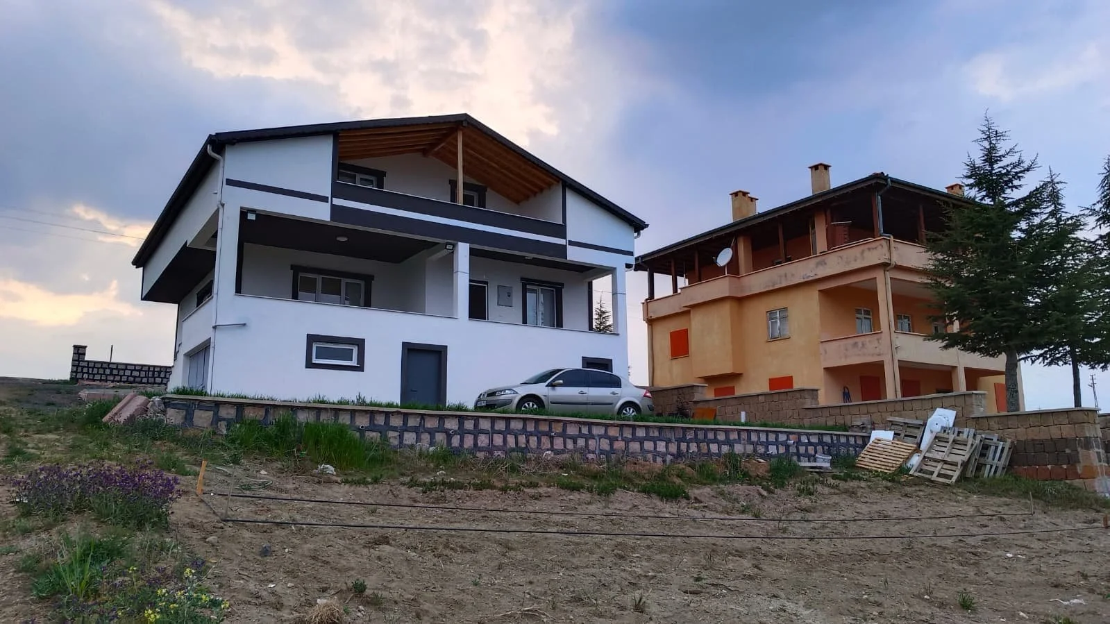 Two houses, one white and one orange, on a sloped lot with a car parked in front. There are trees, building materials, and a partly cloudy sky.