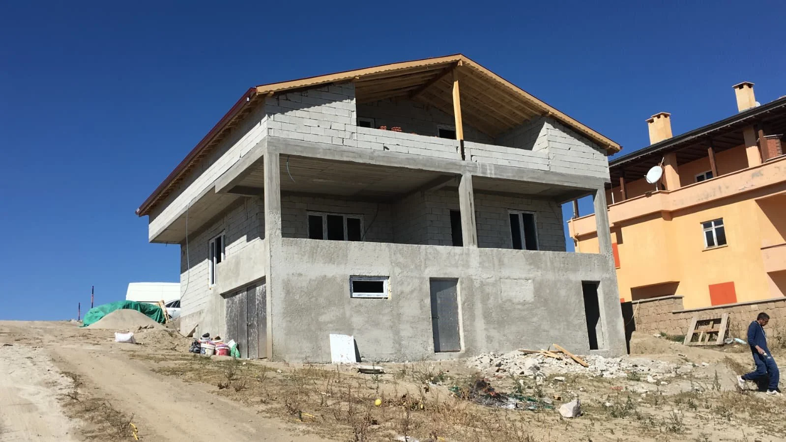 A two-story house under construction with a concrete exterior, located on a dirt lot. Next to it, a multi-story building with an orange facade and satellite dish on the balcony. A man walks on the dirt, and construction materials are scattered around