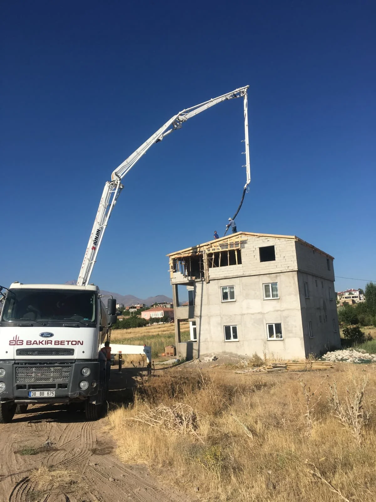 A construction site with a concrete truck pouring cement into the upper floor of a multi-story building under construction. The building is unfinished, with some windows installed and wooden framing visible at the top. The scene takes place in a rura