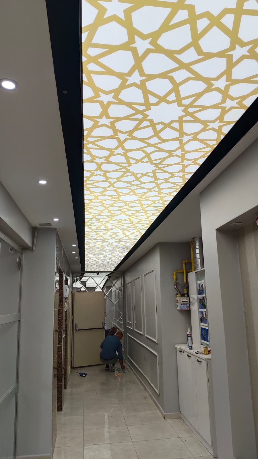 Ceiling with decorative gold geometric pattern LED light panel in a modern interior hallway with two workers installing or repairing.