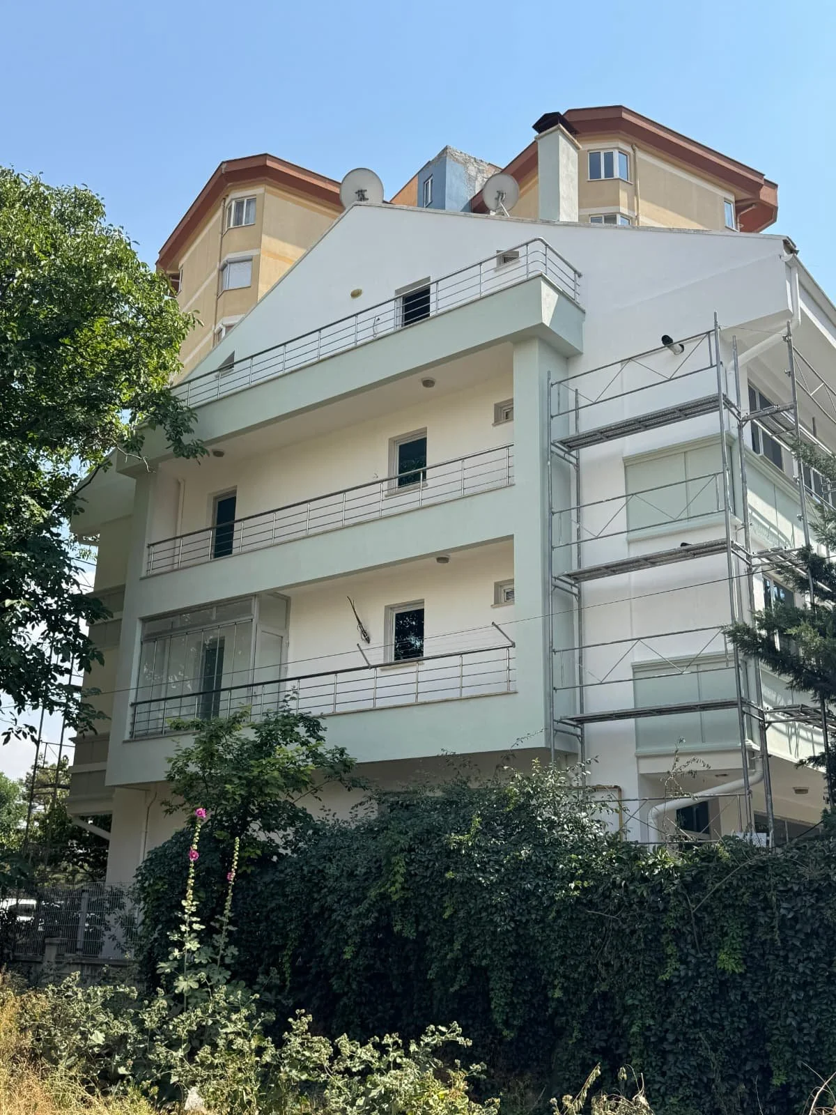 Multi-story residential building with white exterior, metal railings on balconies, and scaffolding on one side, surrounded by trees and greenery.