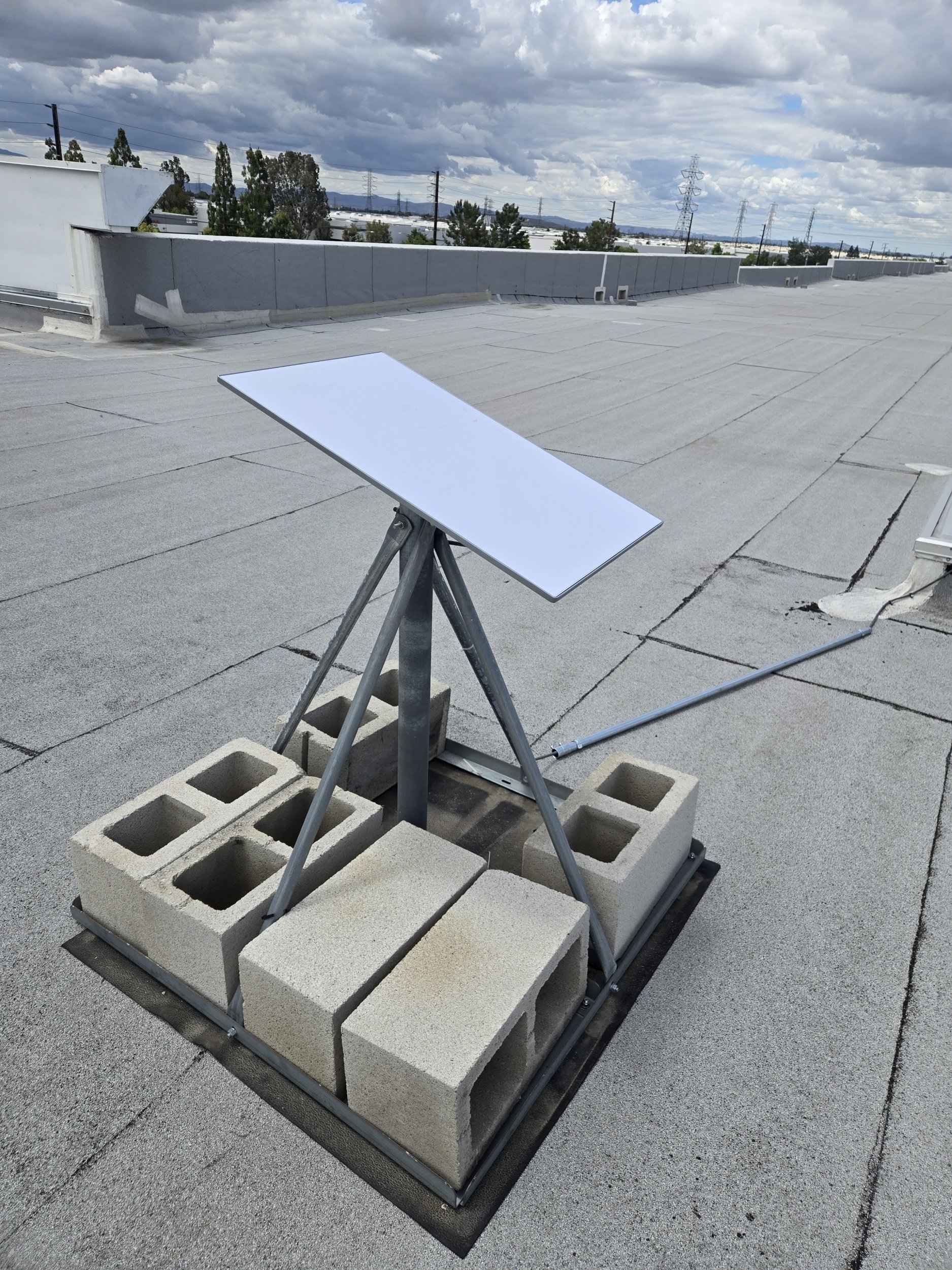 A flat rooftop with cinder blocks arranged in a grid pattern, a solar panel mounted on a tripod, and a row of concrete barriers under cloudy skies.
