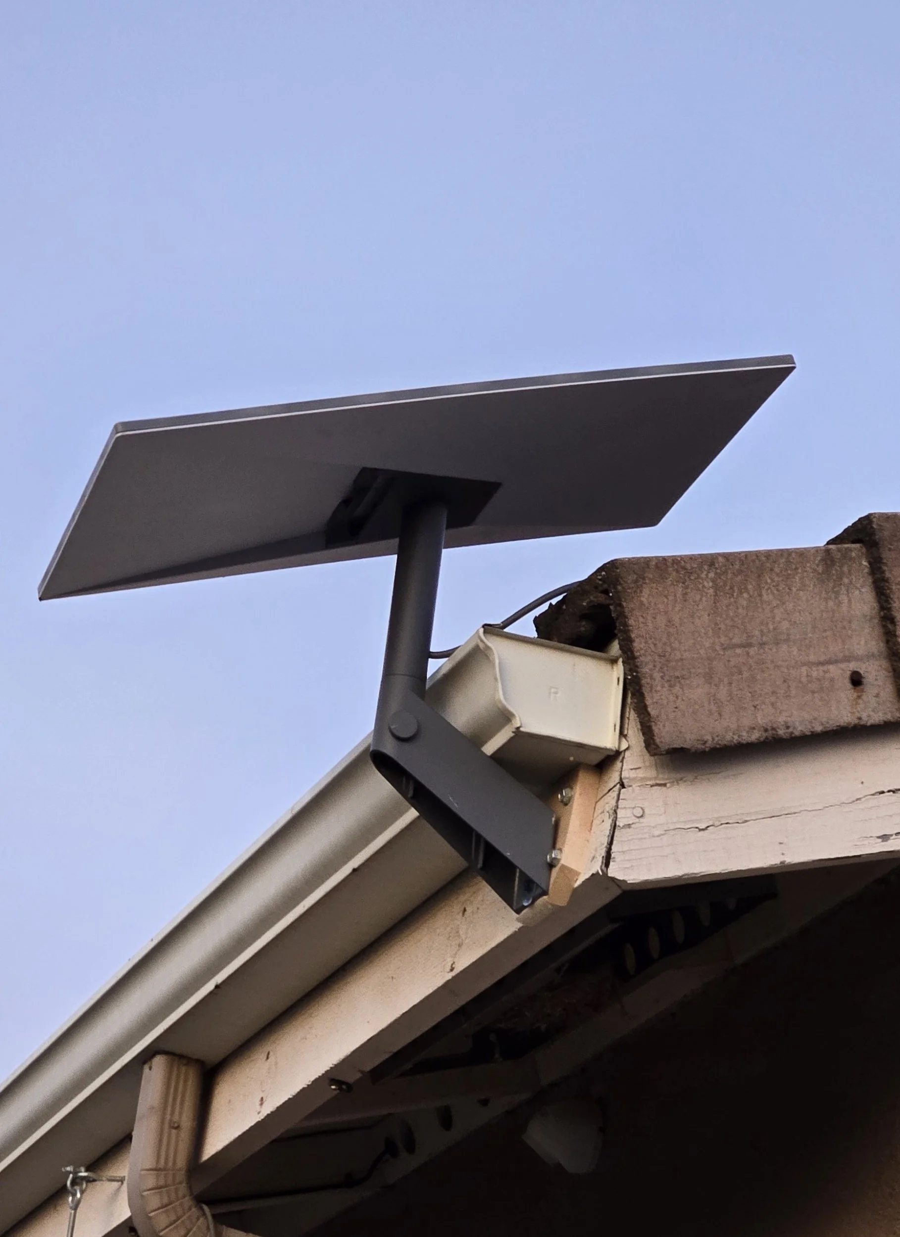 Close-up of a rooftop corner with solar panel, gutter, and electrical box against a clear blue sky.