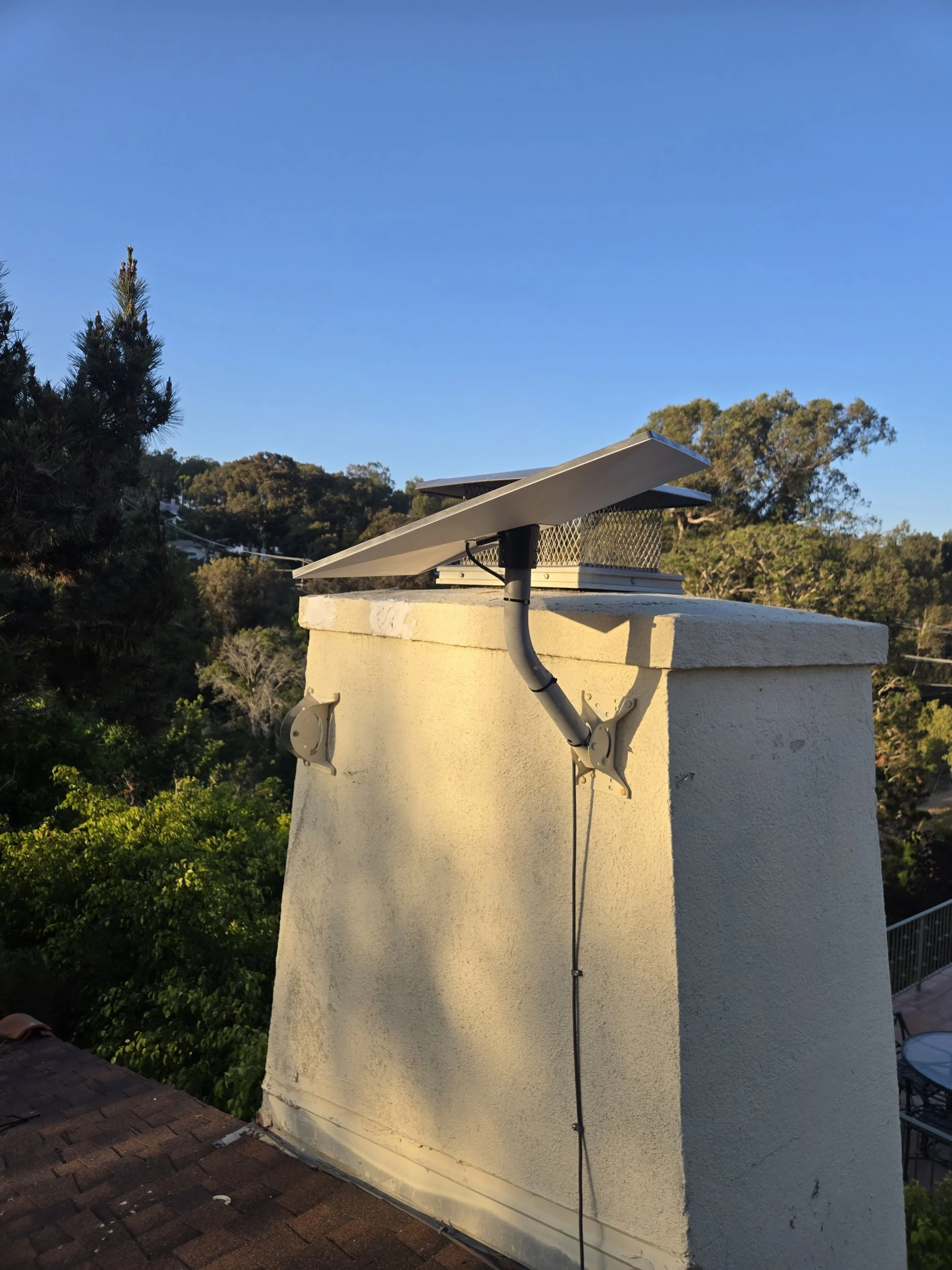 Solar panel installed on a chimney with trees and a clear blue sky in the background.