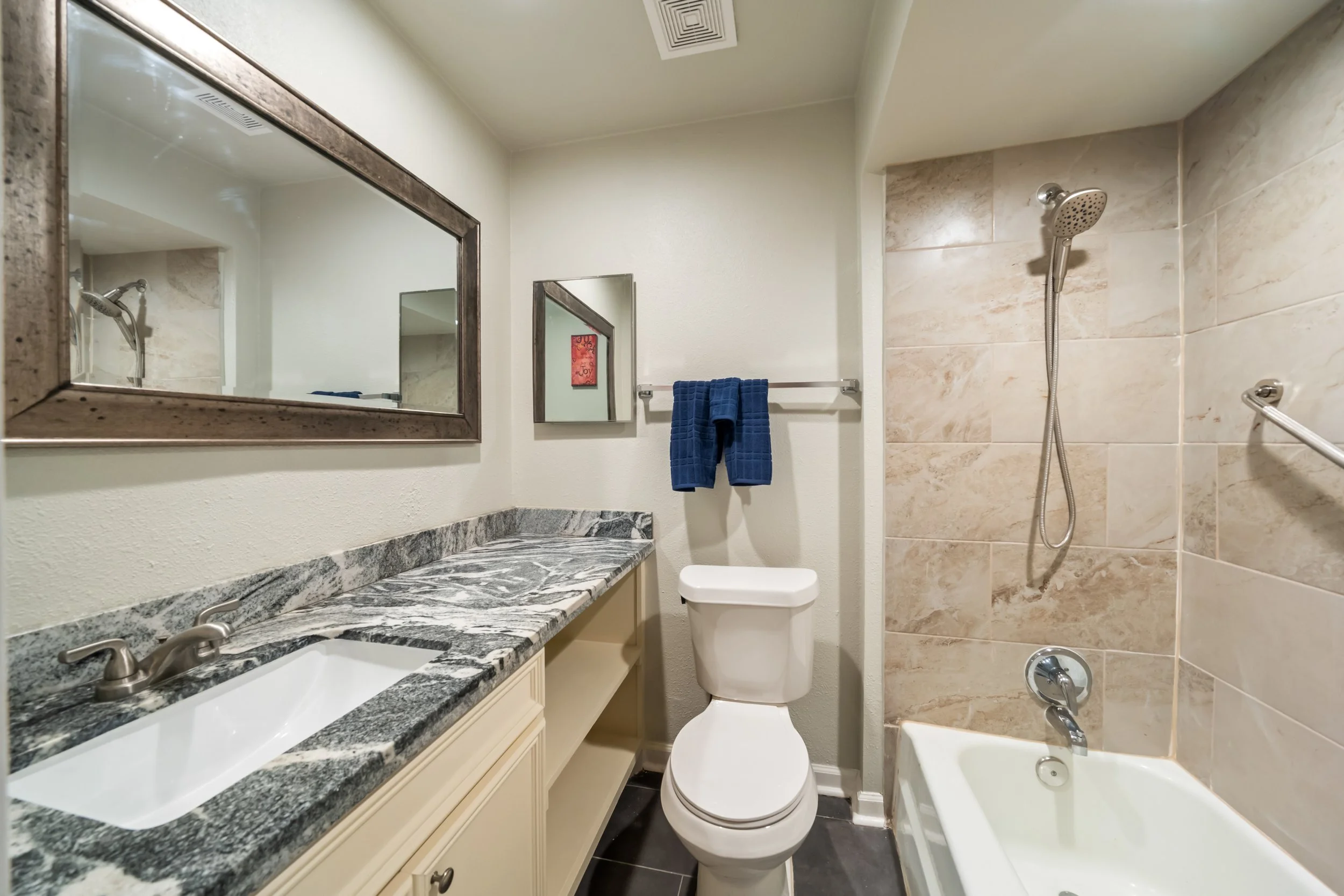 A modern bathroom featuring a granite countertop with dual sinks, a large mirror, a toilet, a shower with a handheld showerhead, and a bathtub. The walls are painted white, and there is a towel rack with a blue towel and small framed artwork on the w