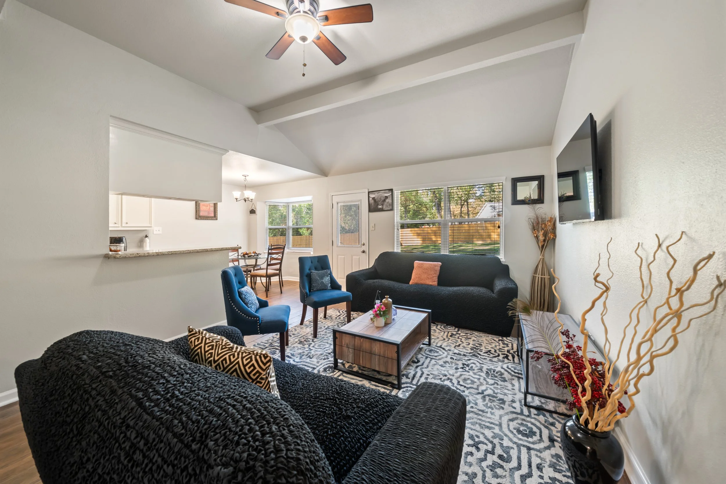 A living room with a black textured sofa, blue chairs, a coffee table with a vase of flowers, a patterned rug, and a wall-mounted TV. There's a ceiling fan, large windows, and a glimpse of the kitchen and dining area.