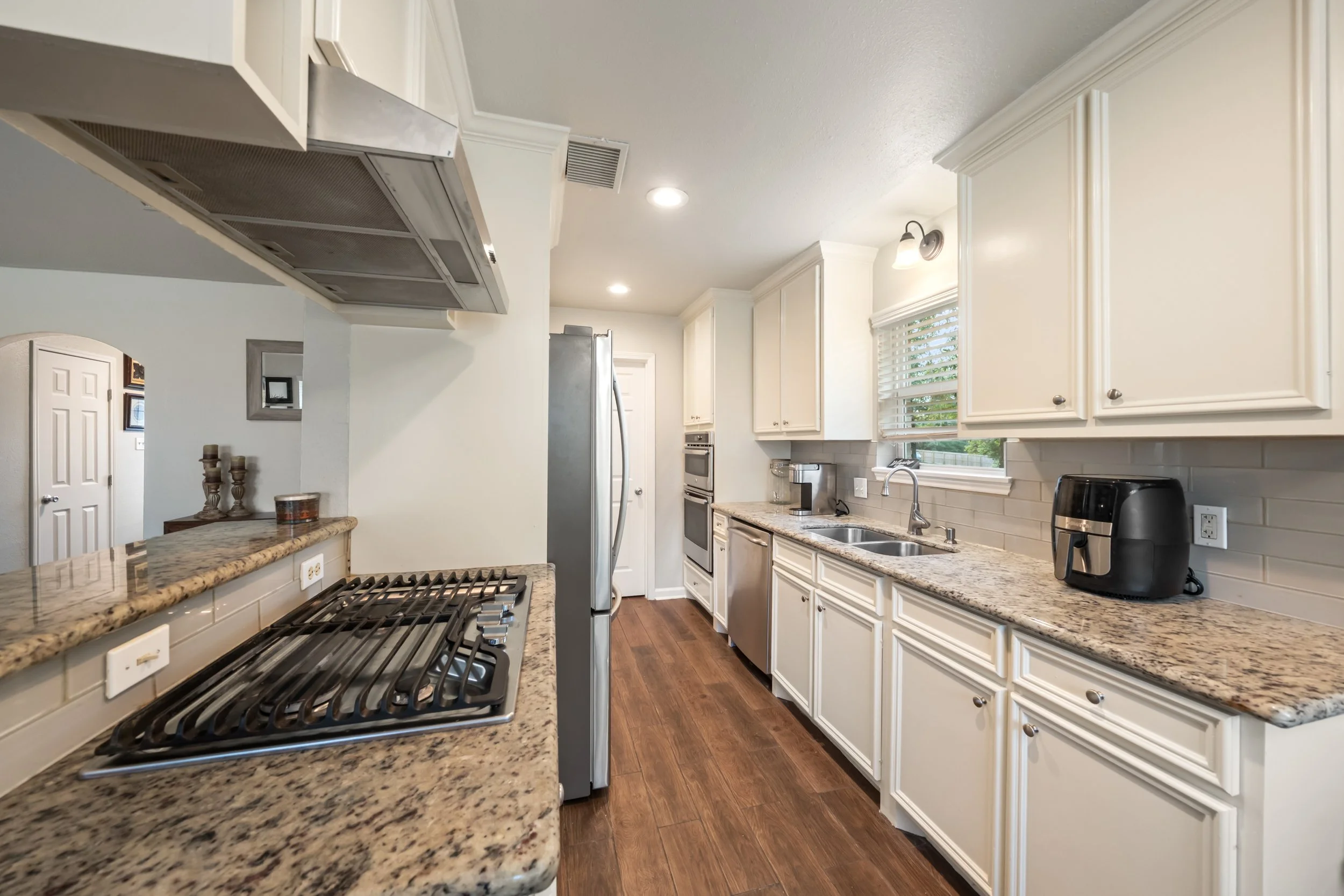 Kitchen with white cabinets, granite countertops, stainless steel appliances including a refrigerator and oven, a double sink under a window, and a black coffee maker on the counter.