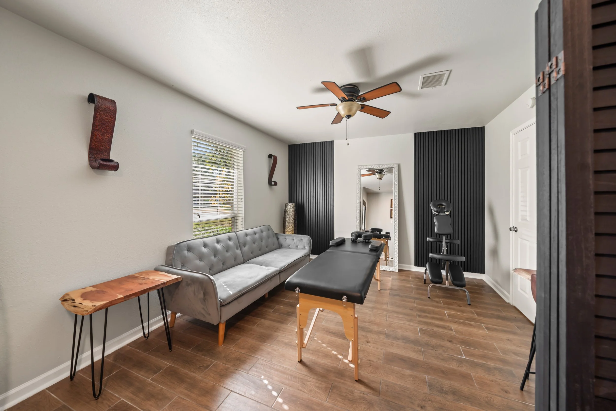 A room with a ceiling fan, a window with blinds, a mirror, a gray tufted sofa, a black massage table, a wooden side table with black metal legs, and black workout equipment, with wood-like tile flooring and neutral-colored walls.