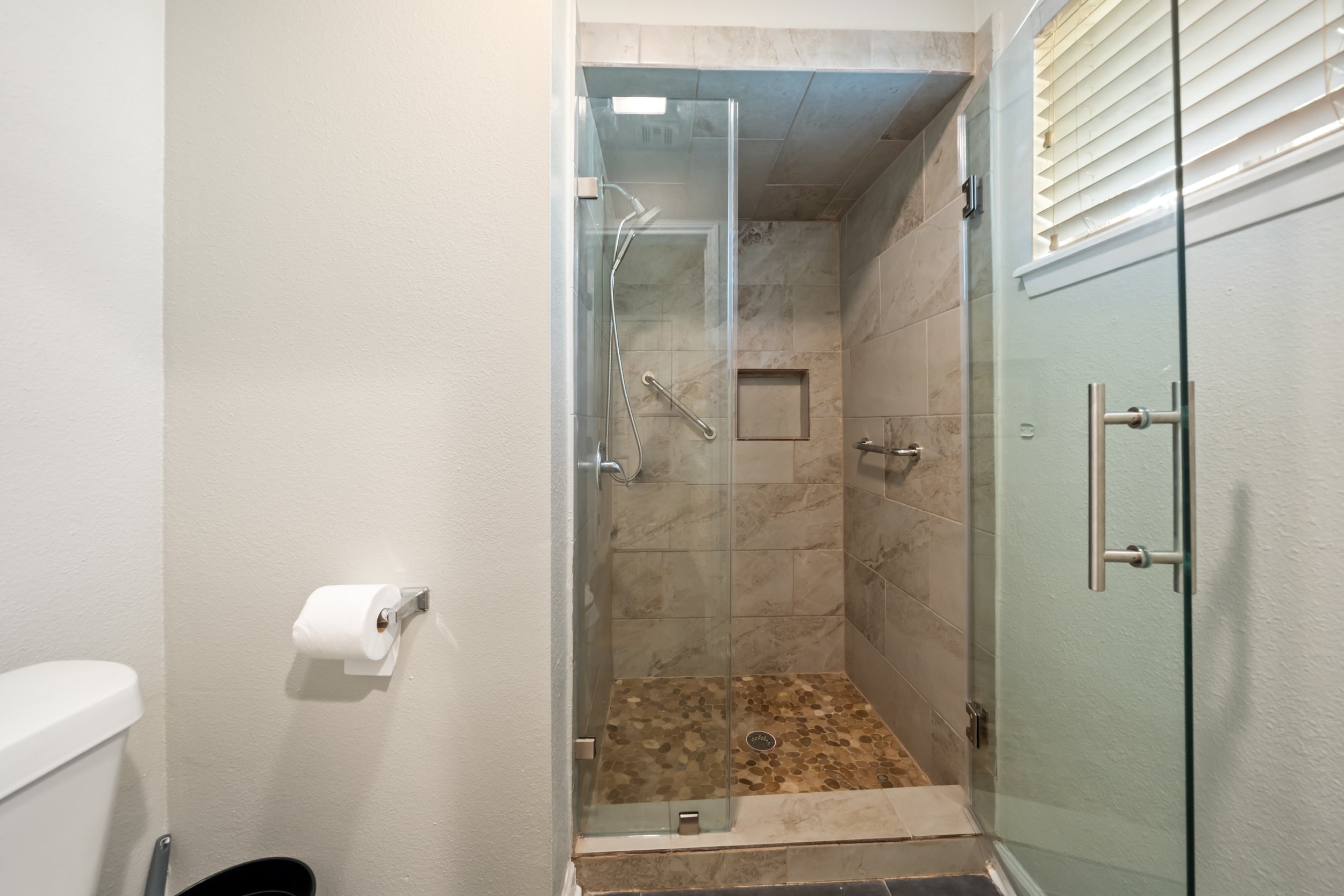 Bathroom with beige tiled shower, glass door, wall-mounted showerhead, and a small window with blinds. Toilet with toilet paper holder and a roll of toilet paper.