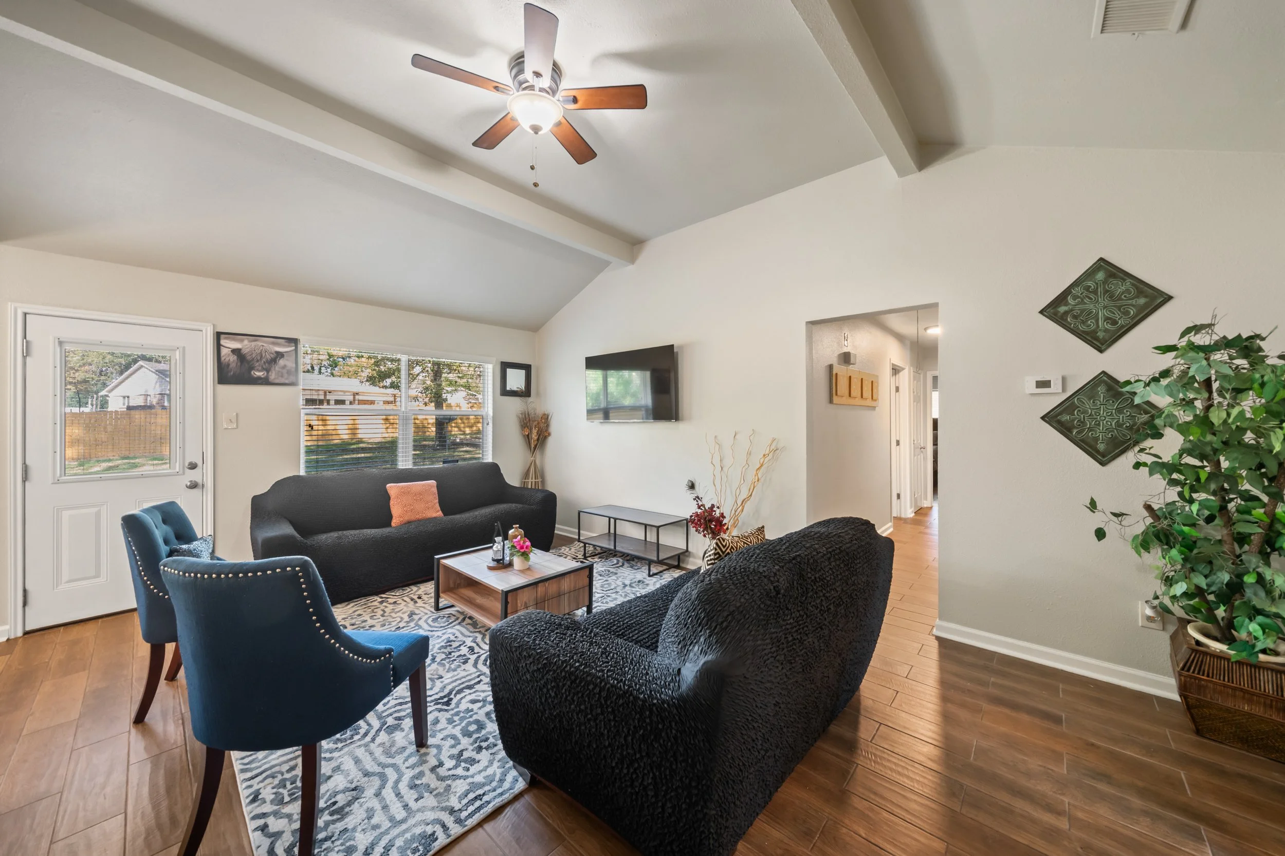 Living room with dark sofa, blue armchairs, a patterned rug, wall decorations, a wall-mounted TV, and large windows
