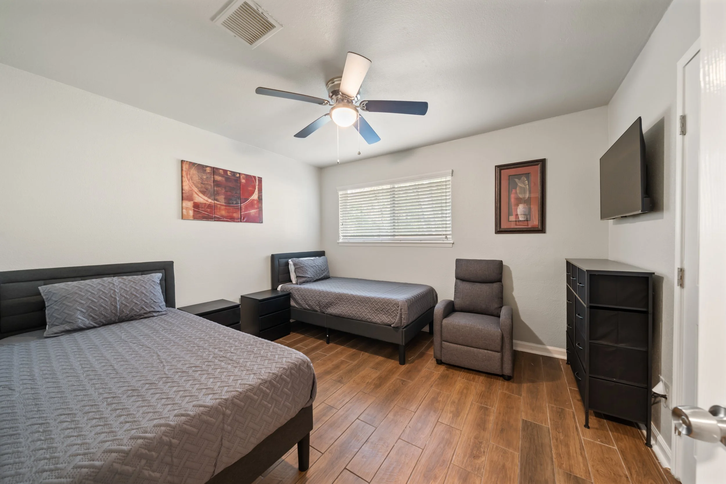 A bedroom with two beds, a ceiling fan, wall art, a small dresser, a flat-screen TV mounted on the wall, and a window with blinds.