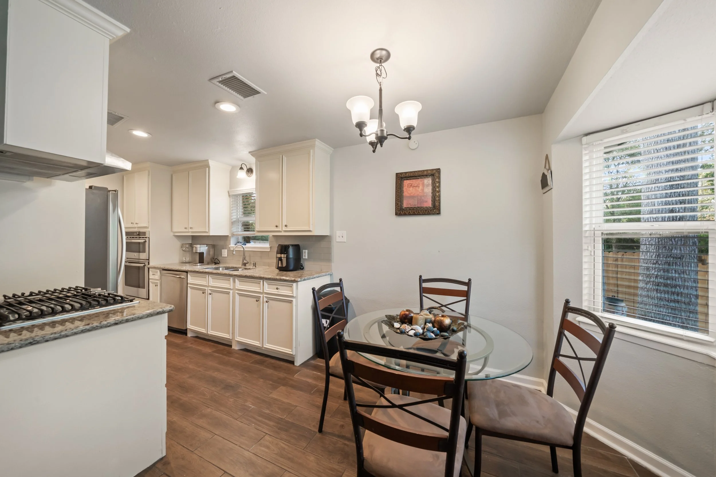 A kitchen with white cabinets, granite countertops, stainless steel appliances, a round glass table with four chairs, a window with blinds, and a chandelier hanging from the ceiling.