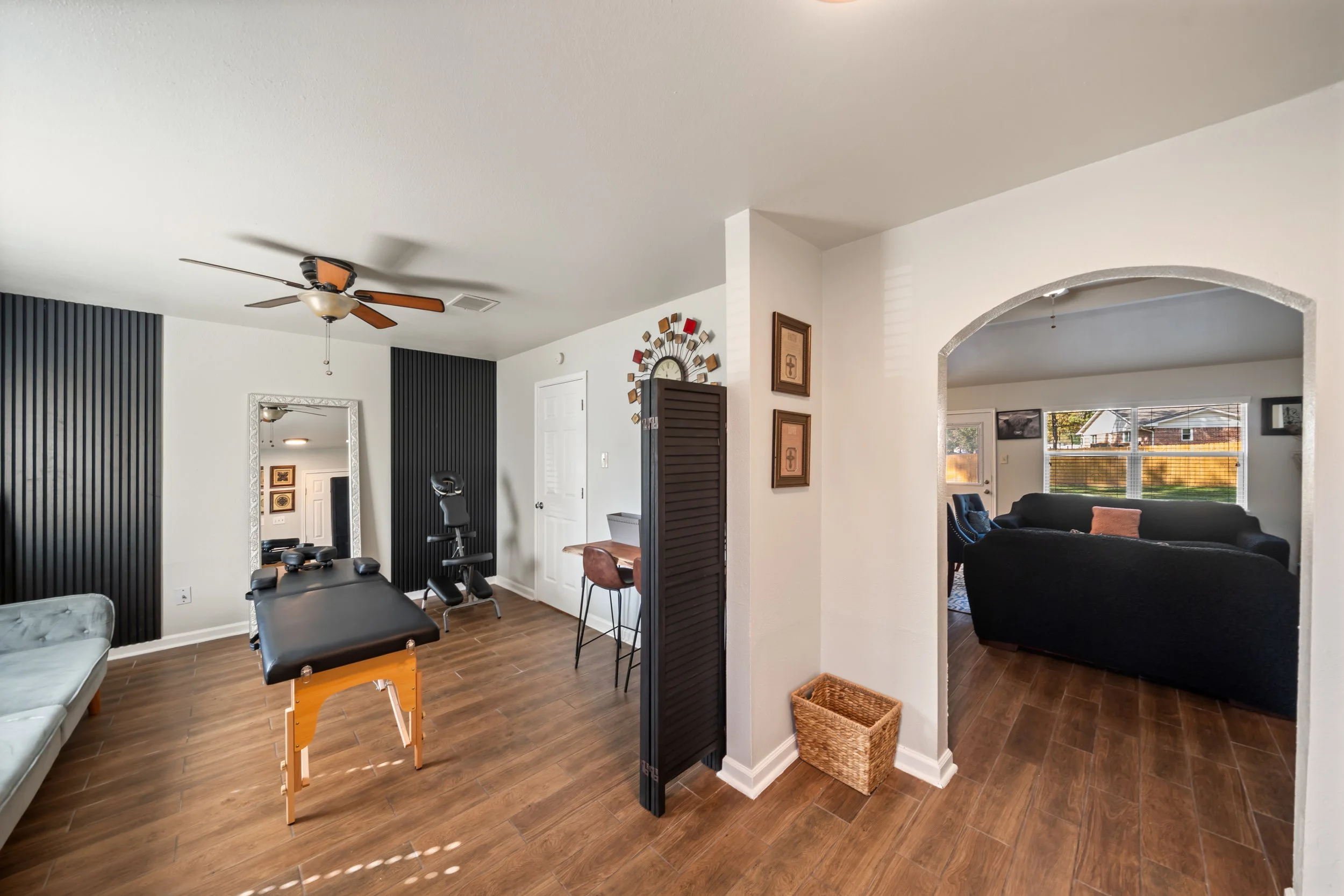 Living room with a ceiling fan, black massage table, wooden flooring, large mirror, black folding door, framed wall art, and two connected rooms with large windows.