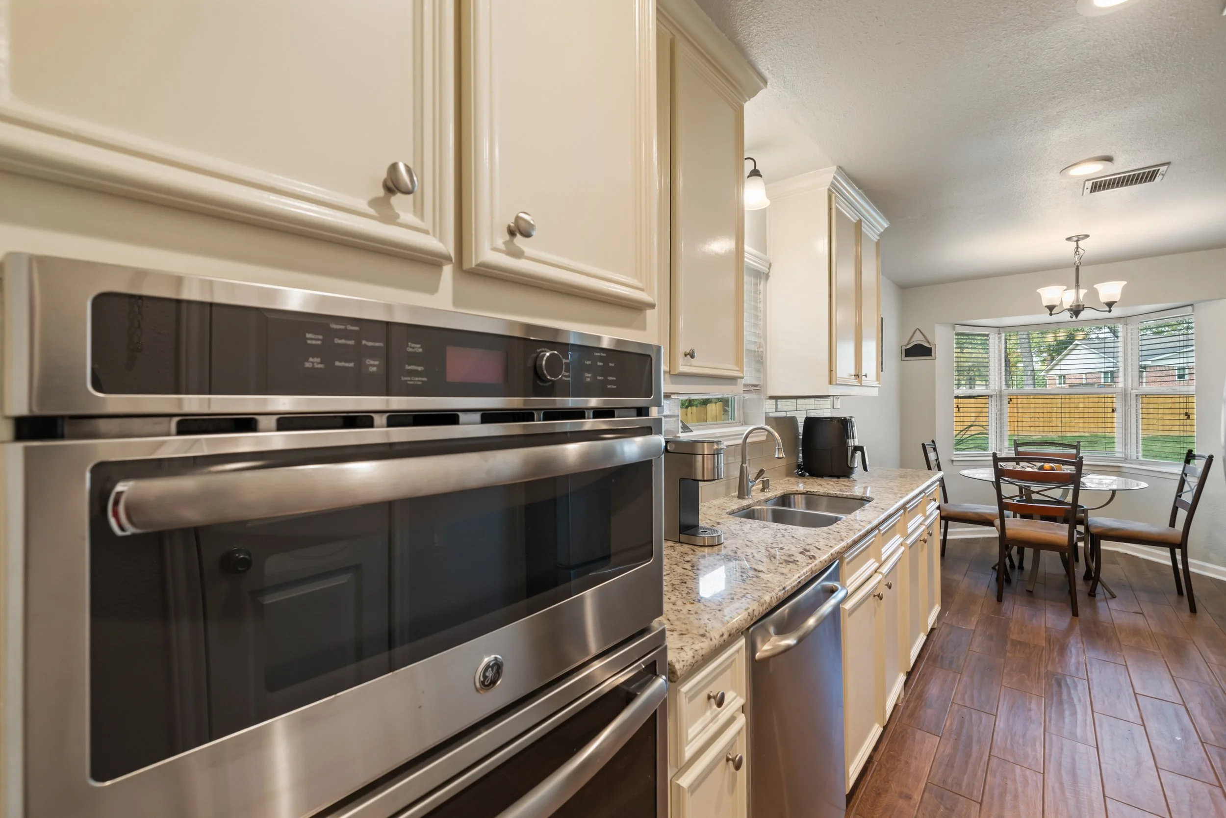 A modern kitchen with beige cabinets, a granite countertop, and a stainless steel oven. There is a double sink, black appliances, a round dining table with chairs, and large windows with blinds overlooking a backyard.