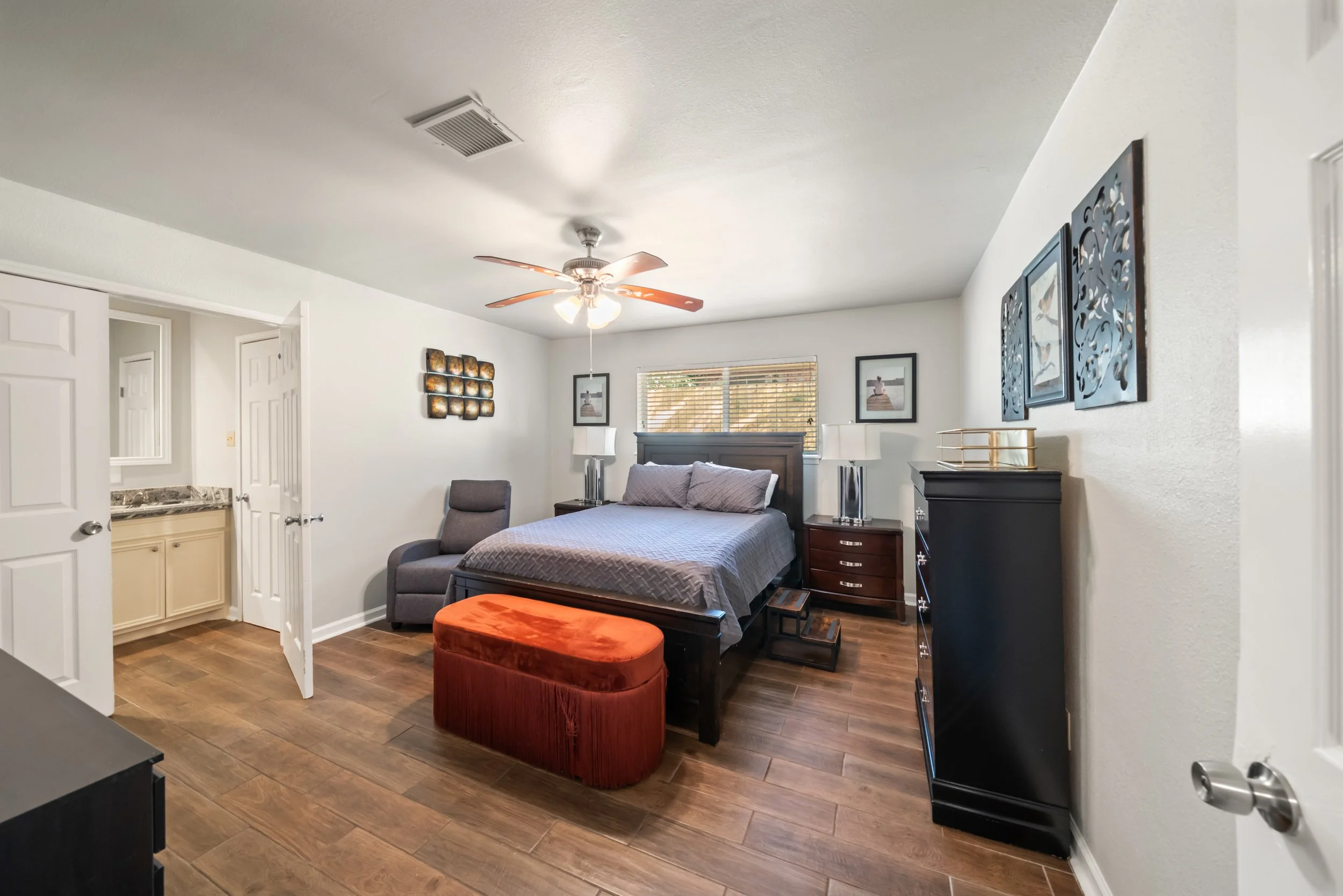 A bedroom with a bed, nightstands, lamps, a sitting chair, and wall decorations, with a window, hardwood floor, ceiling fan, and an open door.