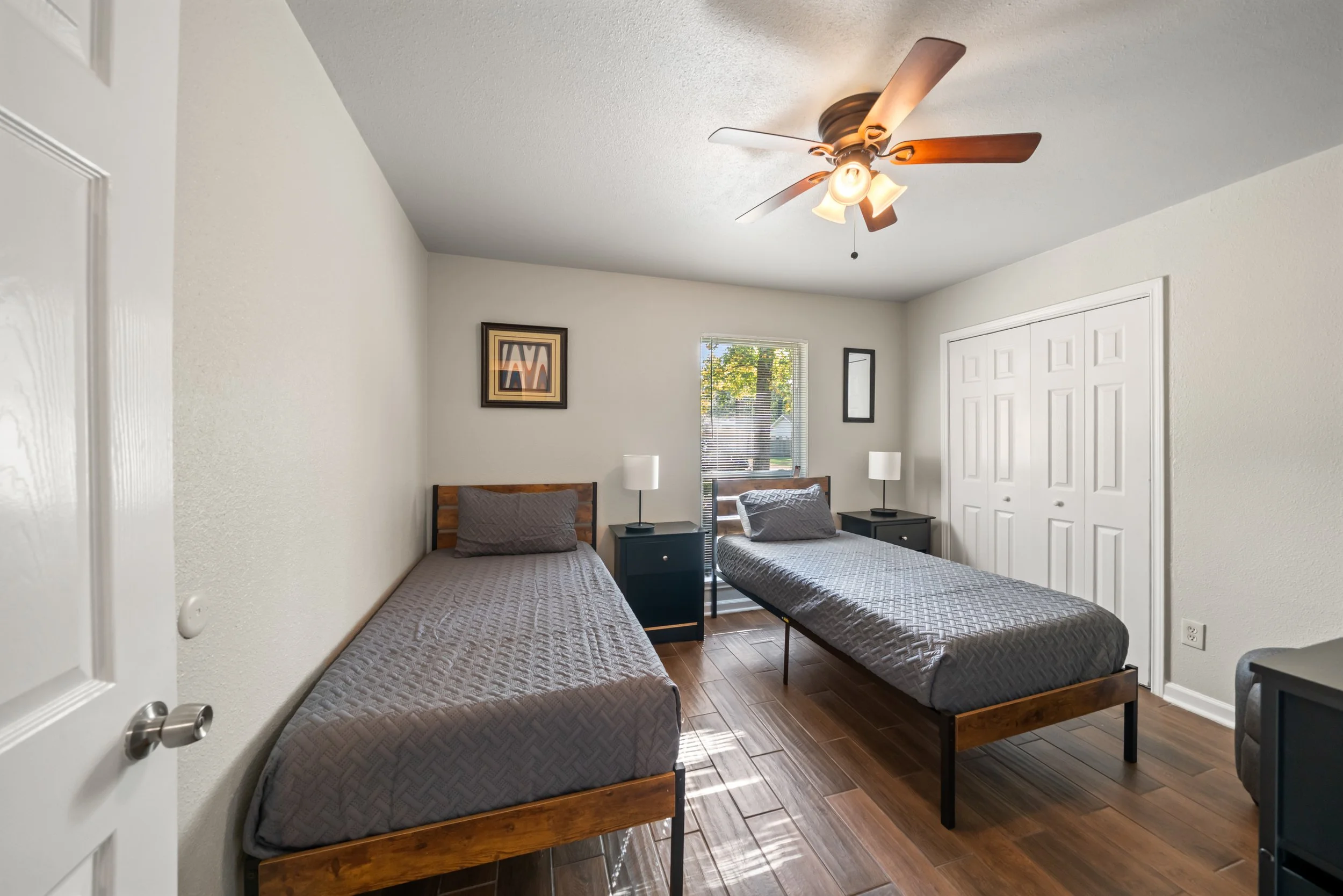 A bedroom with two single beds, each with a gray quilted mattress, placed parallel to each other. Between the beds are black nightstands, each with a white table lamp. A window with blinds showing trees outside is on the wall behind the beds. A frame