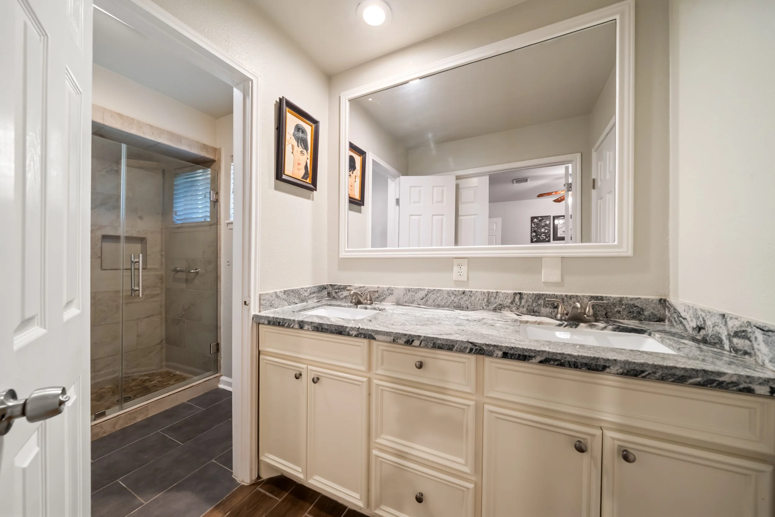 Bathroom vanity with marble countertop and a large mirror, a double sink, and framed artwork on the wall. Visible shower area with glass door and tiled walls.