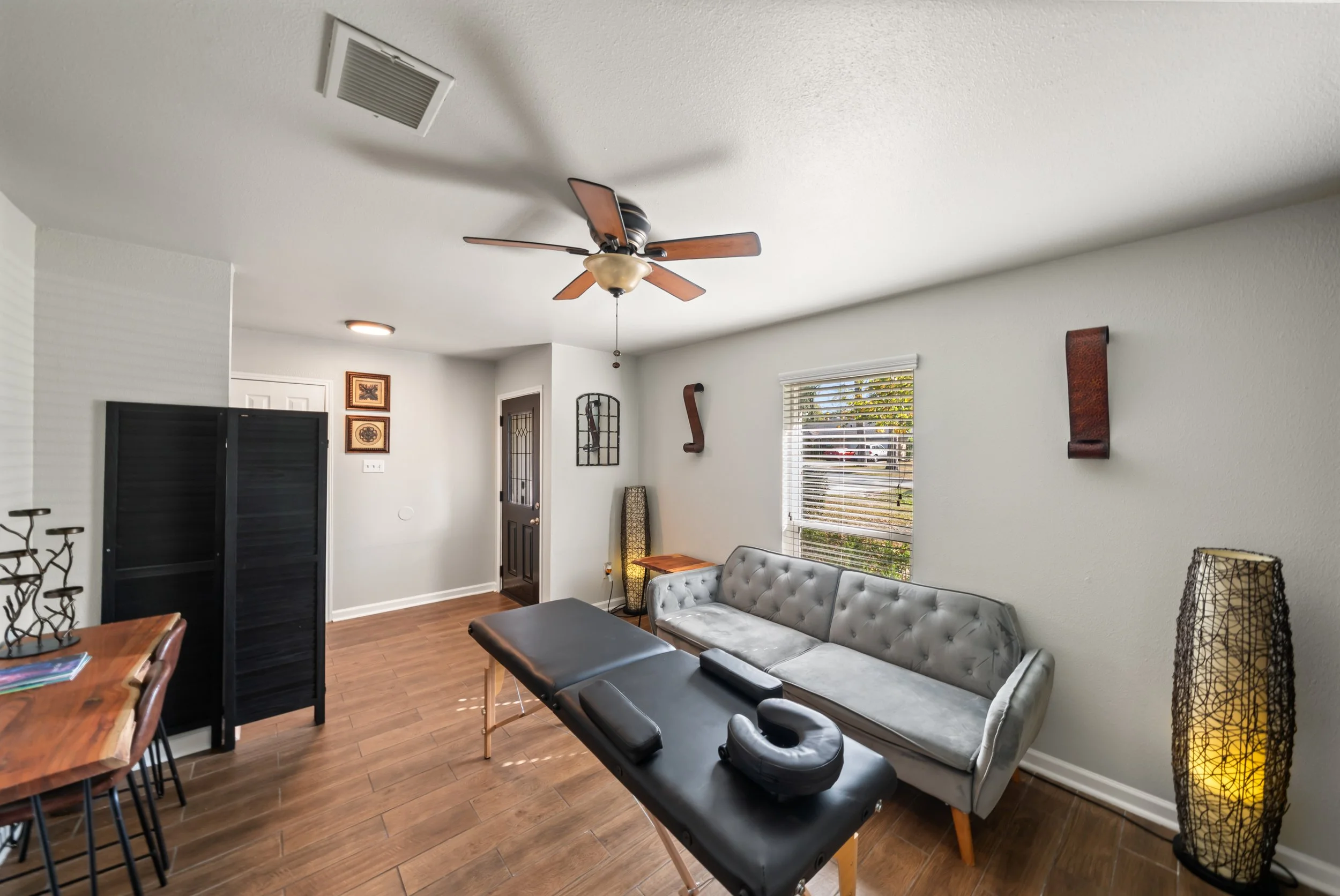 Living room with a ceiling fan, a gray tufted sofa, a massage table, and a window with blinds.