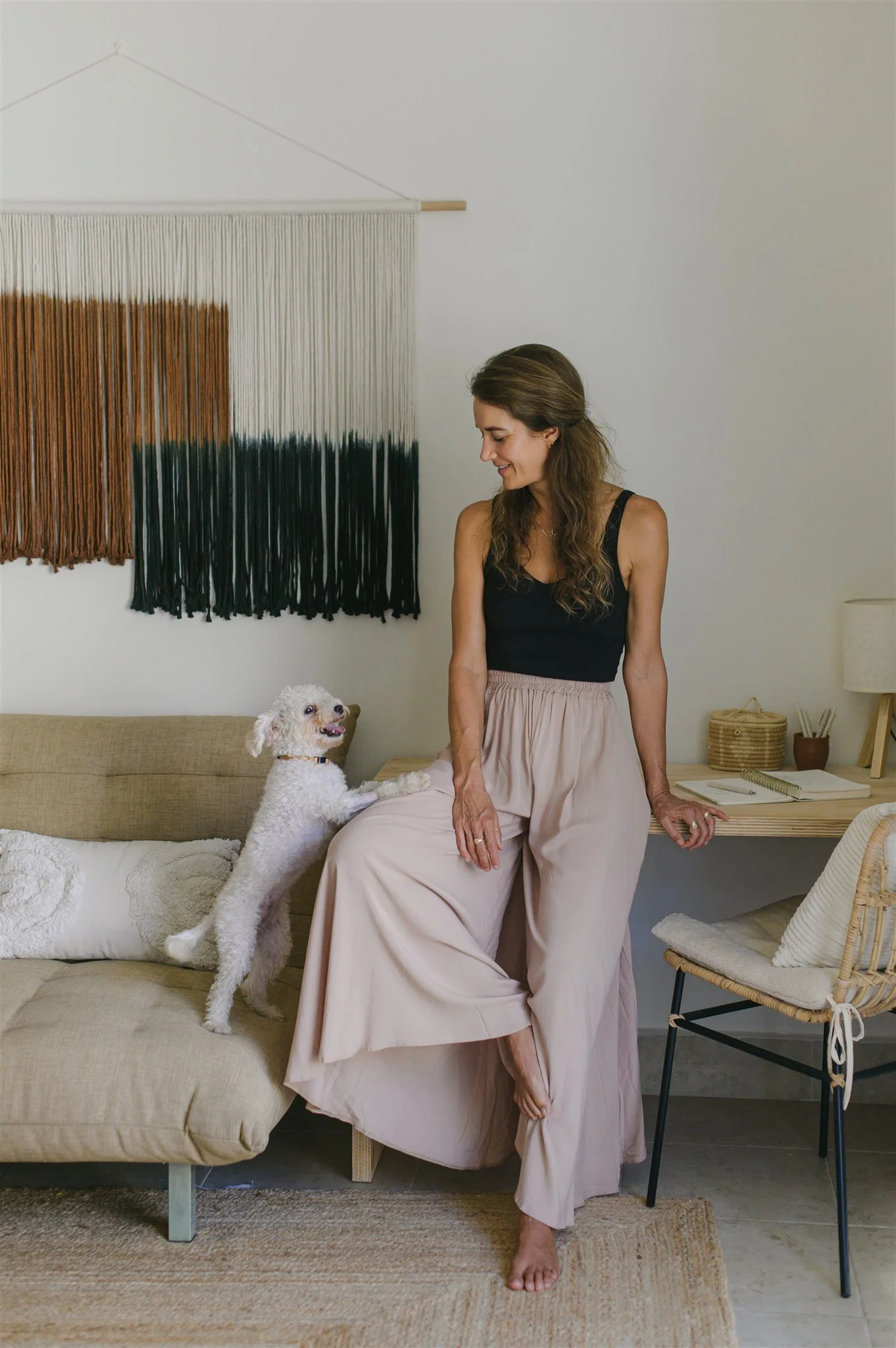 A woman in a black tank top and flowing beige pants is sitting on the edge of a beige sofa, smiling and looking at her white poodle standing on its hind legs with paws on her lap. The setting is a minimally decorated room with a woven wall hanging and a wooden desk with a notebook and wicker baskets.