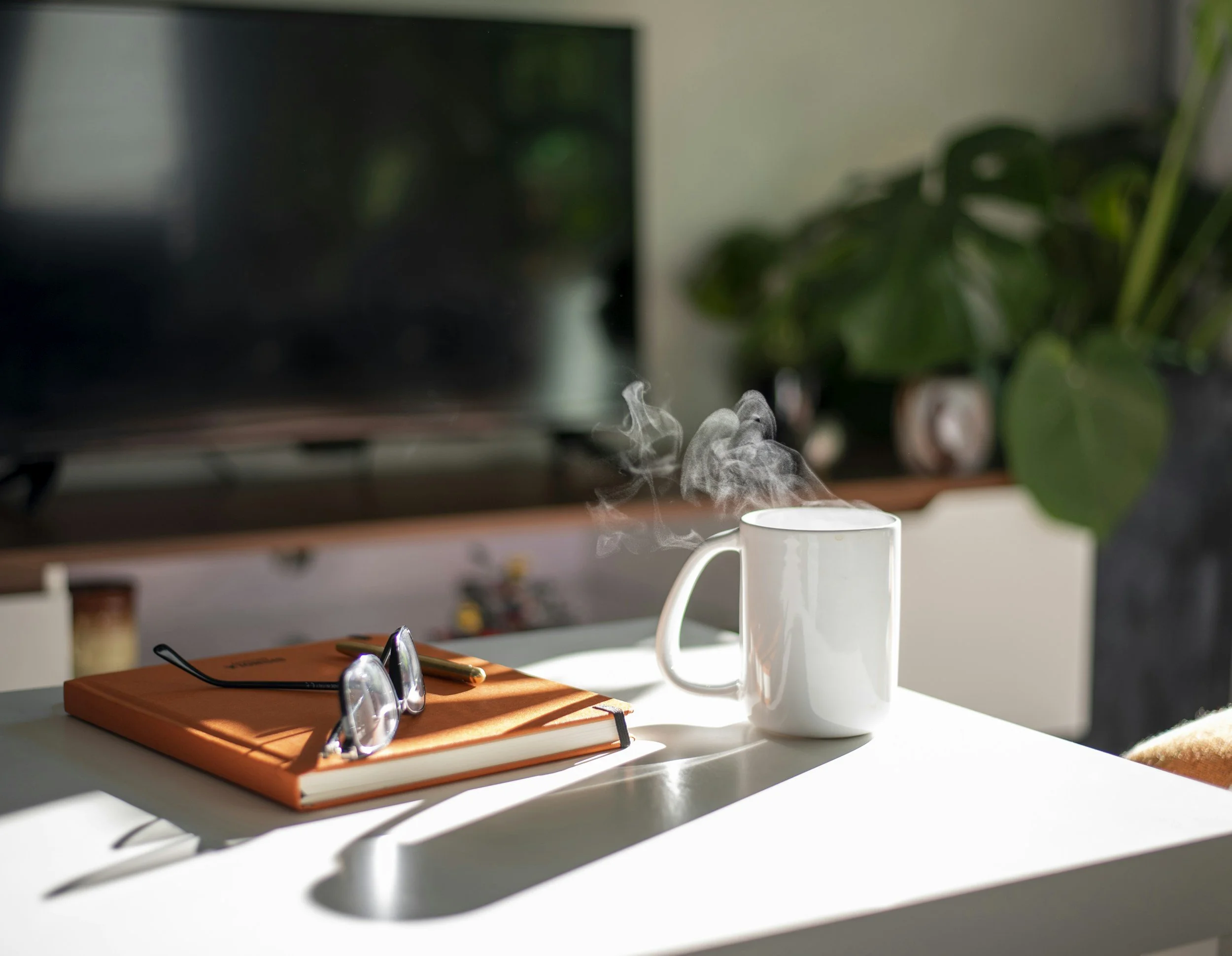 A white coffee mug with steam rising on a white desk, next to a closed orange notebook with eyeglasses resting on top, in a sunlit room with a blurred television and a green plant in the background.