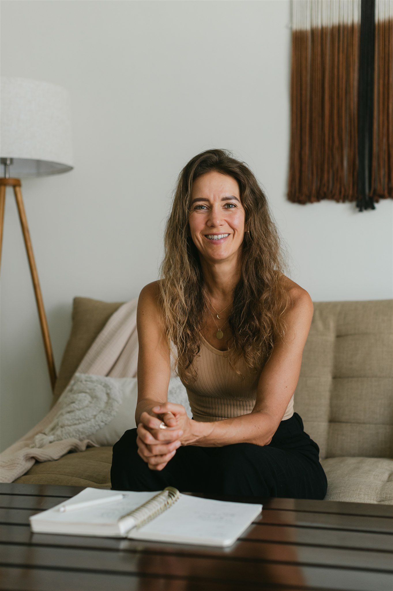 A smiling woman with long wavy brown hair, wearing a beige sleeveless top, sitting on a beige couch in a room with a white wall, a wooden table with an open notebook and a pen, and a lamp with a white shade and wooden tripod legs.