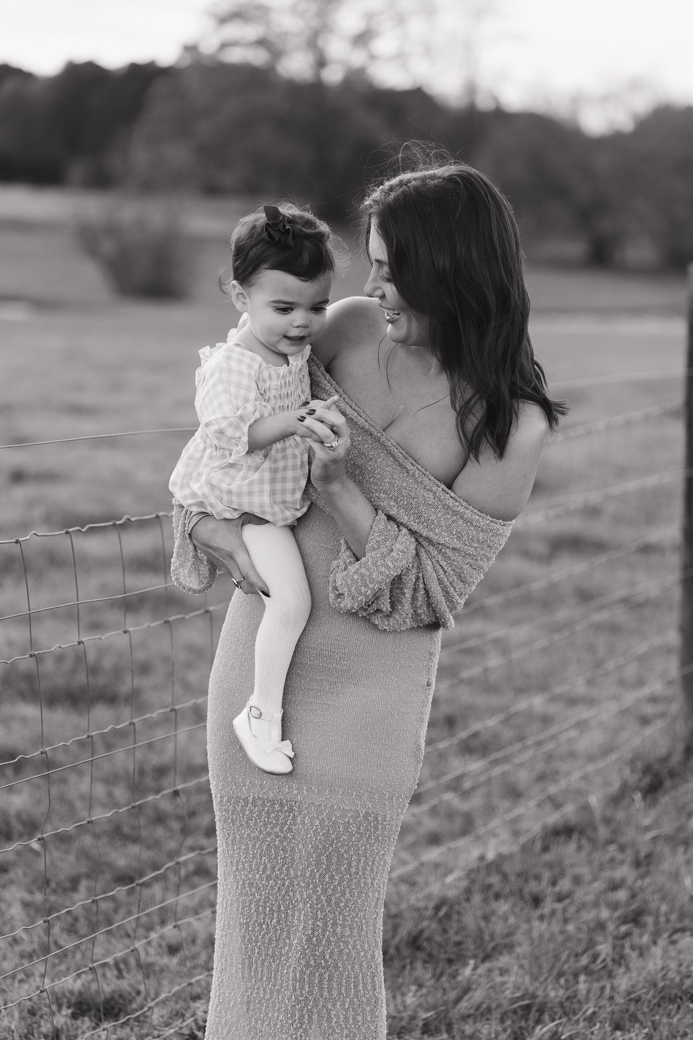 A woman holding a young girl outdoors near a fence with a grassy field and trees in the background, both smiling and looking at each other.