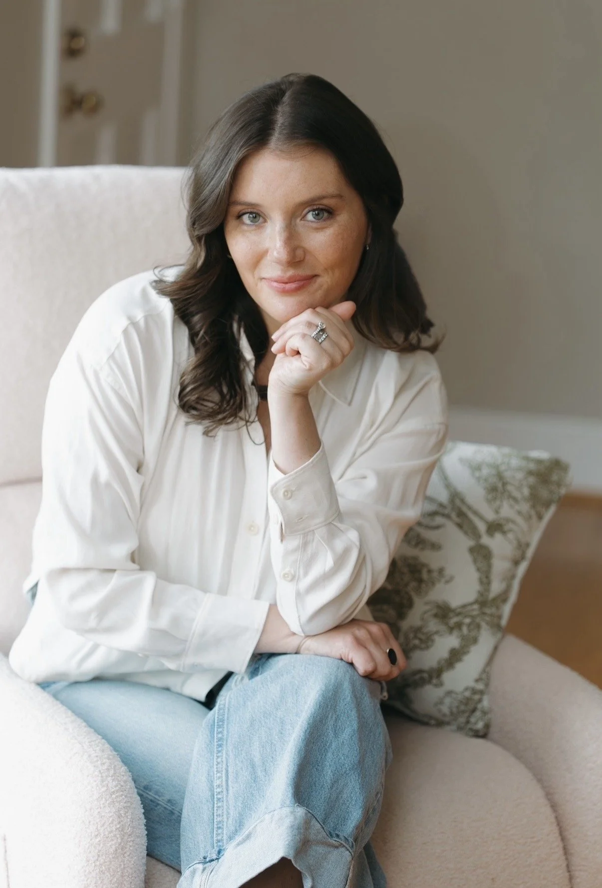 A woman with wavy dark hair and blue eyes sitting on a beige couch, wearing a cream-colored button-up shirt and light blue jeans, smiling softly at the camera with her right hand resting on her chin and her left hand on her knee.