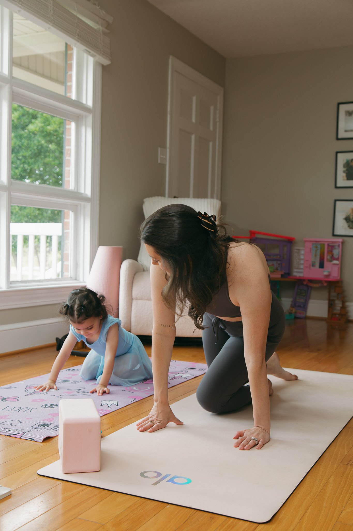 A woman and a young girl practicing yoga together on exercise mats in a bright living room, with large windows and children's toys in the background.