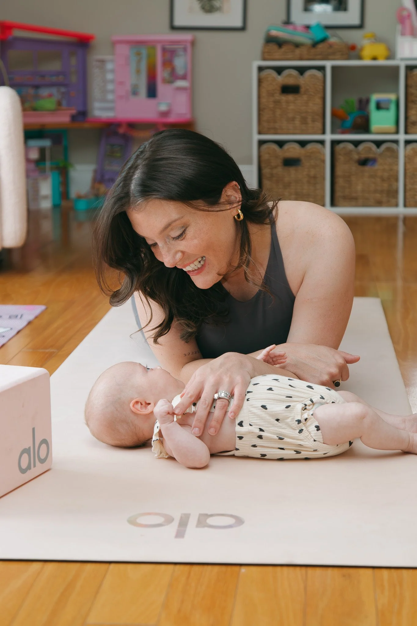 A woman with dark wavy hair and earrings is smiling as she interacts with a baby lying on a beige play mat in a playroom. The baby is dressed in a polka dot outfit and looking up at the woman. Shelves with toys and books are visible in the background.