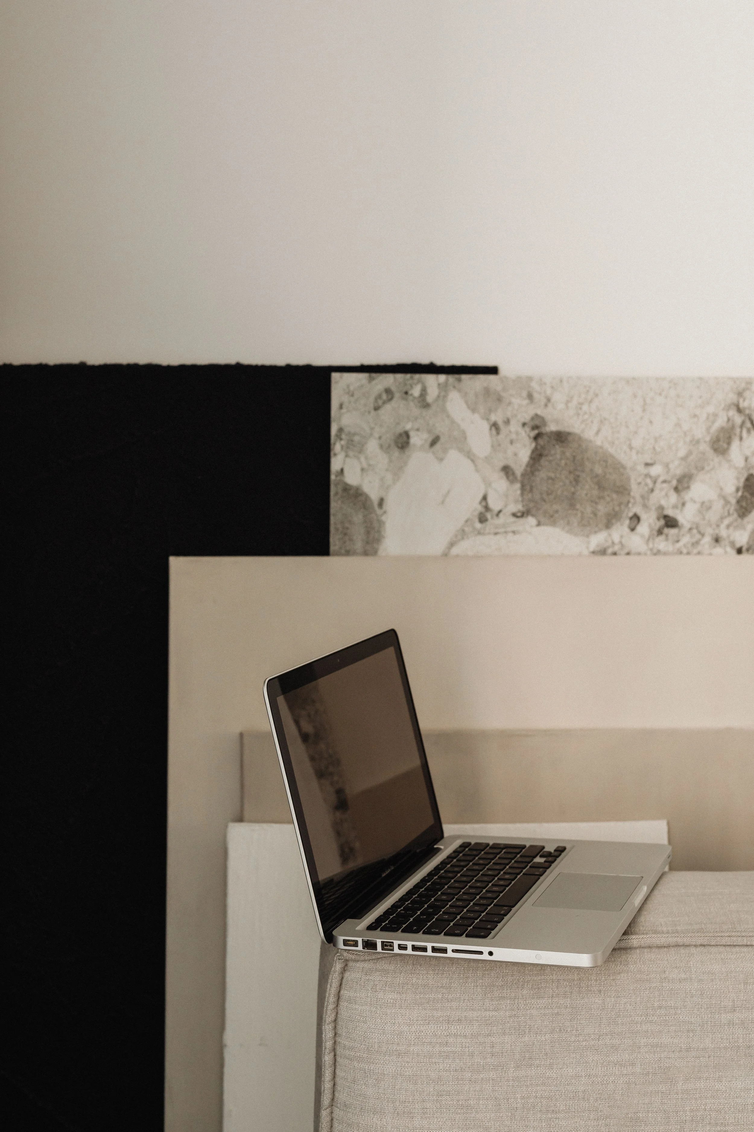 A silver laptop resting on a beige cushioned surface with a black screen and keyboard. In the background, there is a white and gray abstract marbled rectangular object.