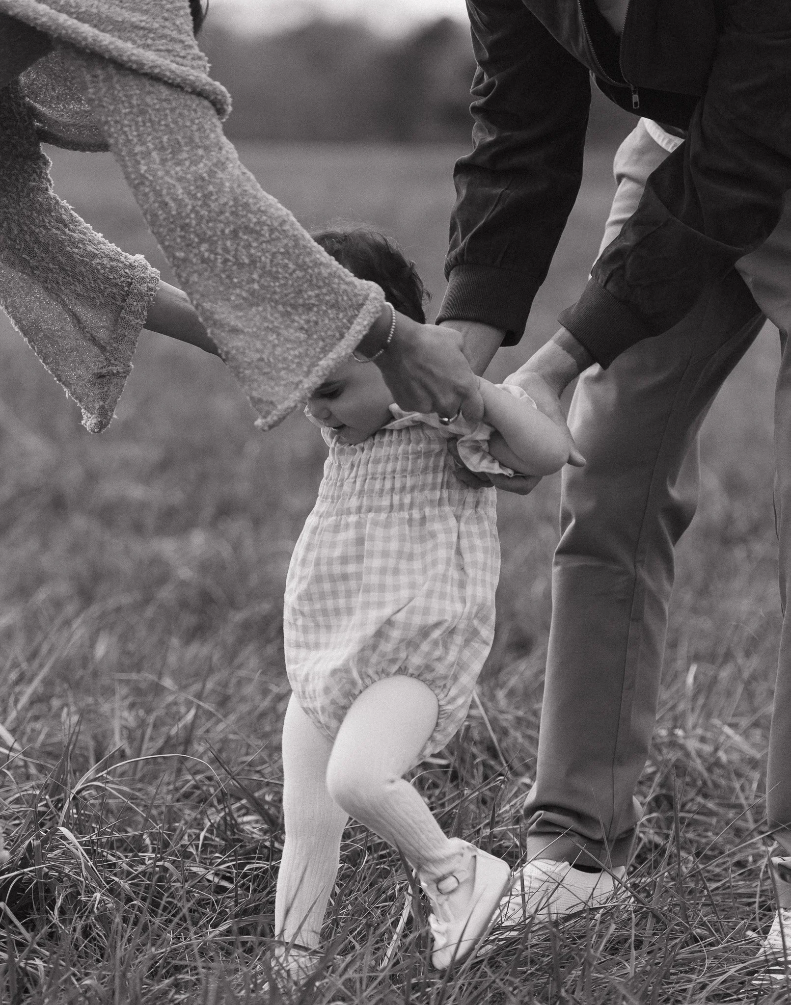 A young child and two adults holding hands while walking through tall grass in an outdoor setting, black and white photo.