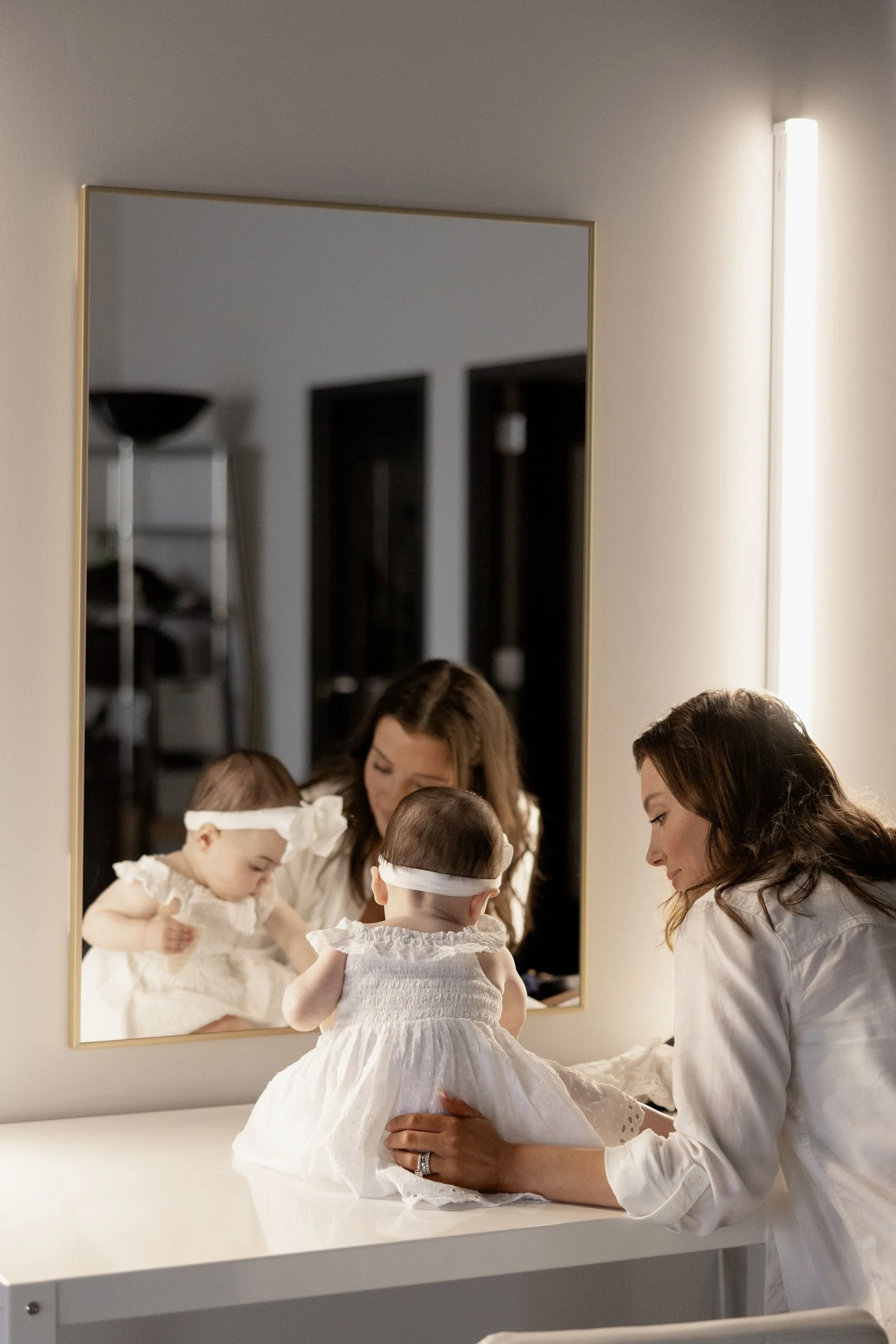 A woman dressed in white looks at two young girls wearing matching white dresses and headbands, with her reflection visible in a mirror on the wall as they prepare for a special occasion.