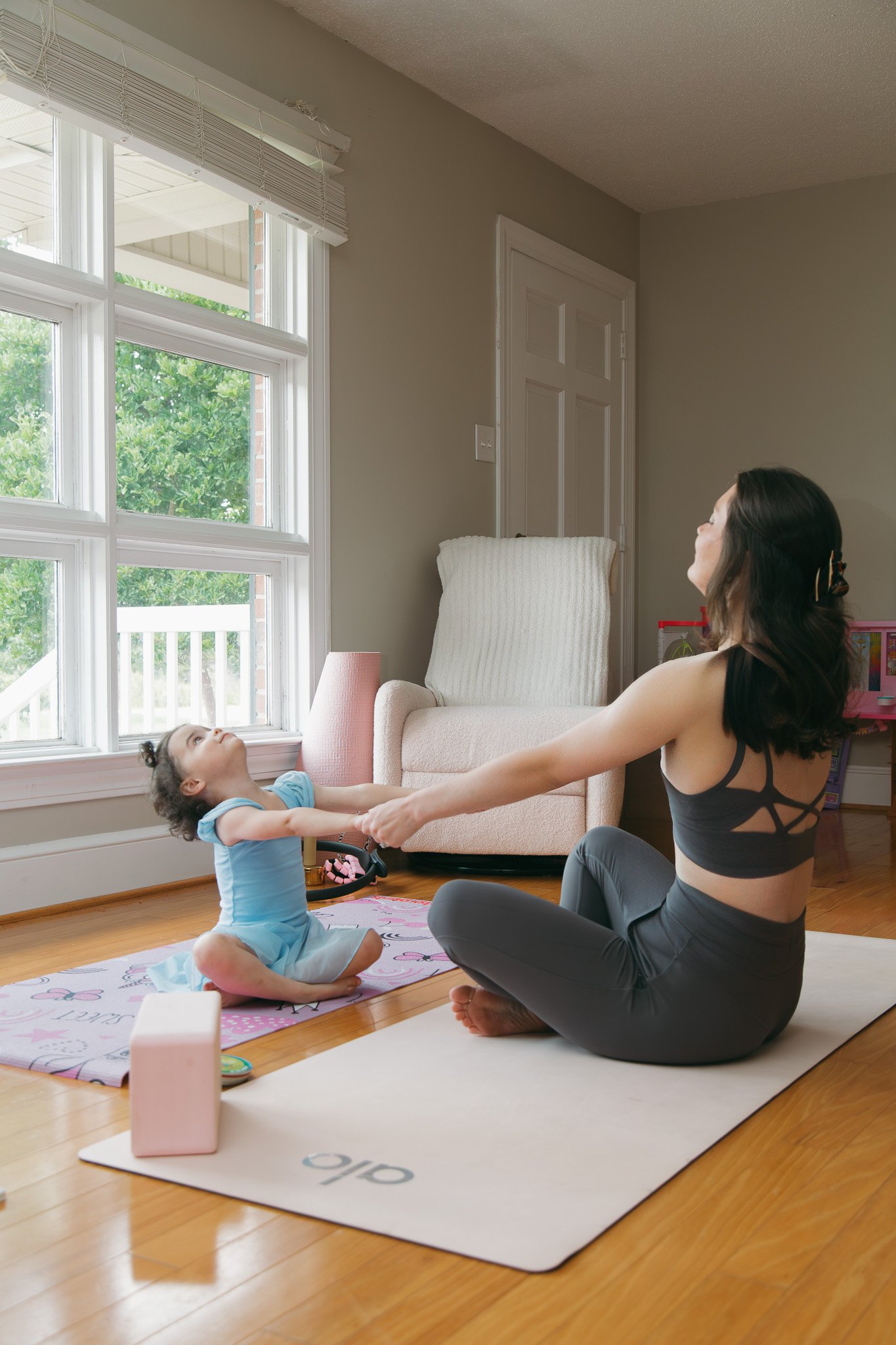 A woman and a young girl are practicing yoga together in a living room. The girl is sitting on the floor with a yoga mat, holding the woman's hands while they perform a partner stretch. The woman, sitting cross-legged, is smiling and looking at the girl. Large windows with blinds let in natural light, and there is a cream-colored armchair and a pink toy storage container in the background.