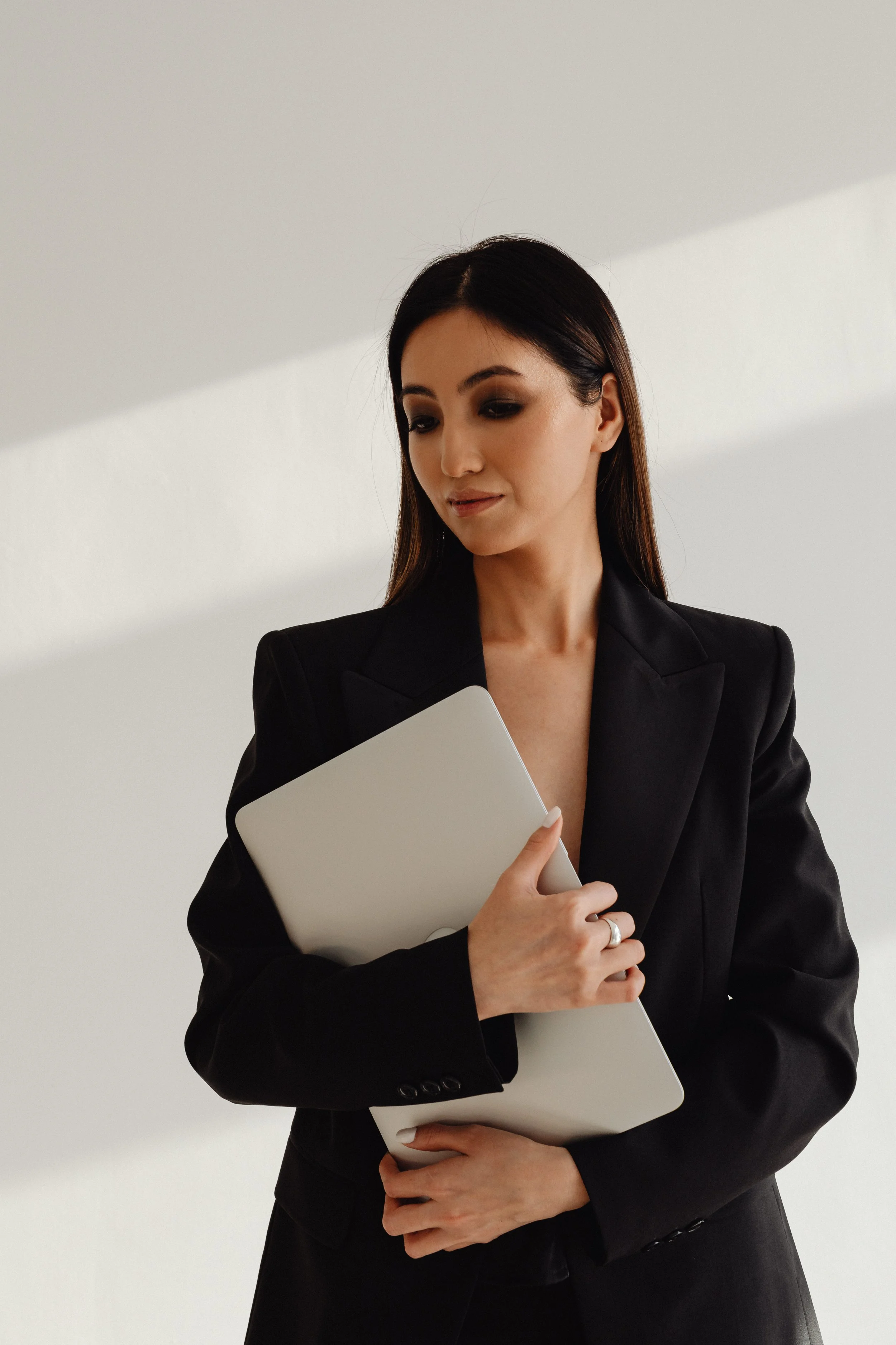 A woman with dark hair styled straight, wearing a black blazer, holding a closed silver laptop, standing against a plain white background.