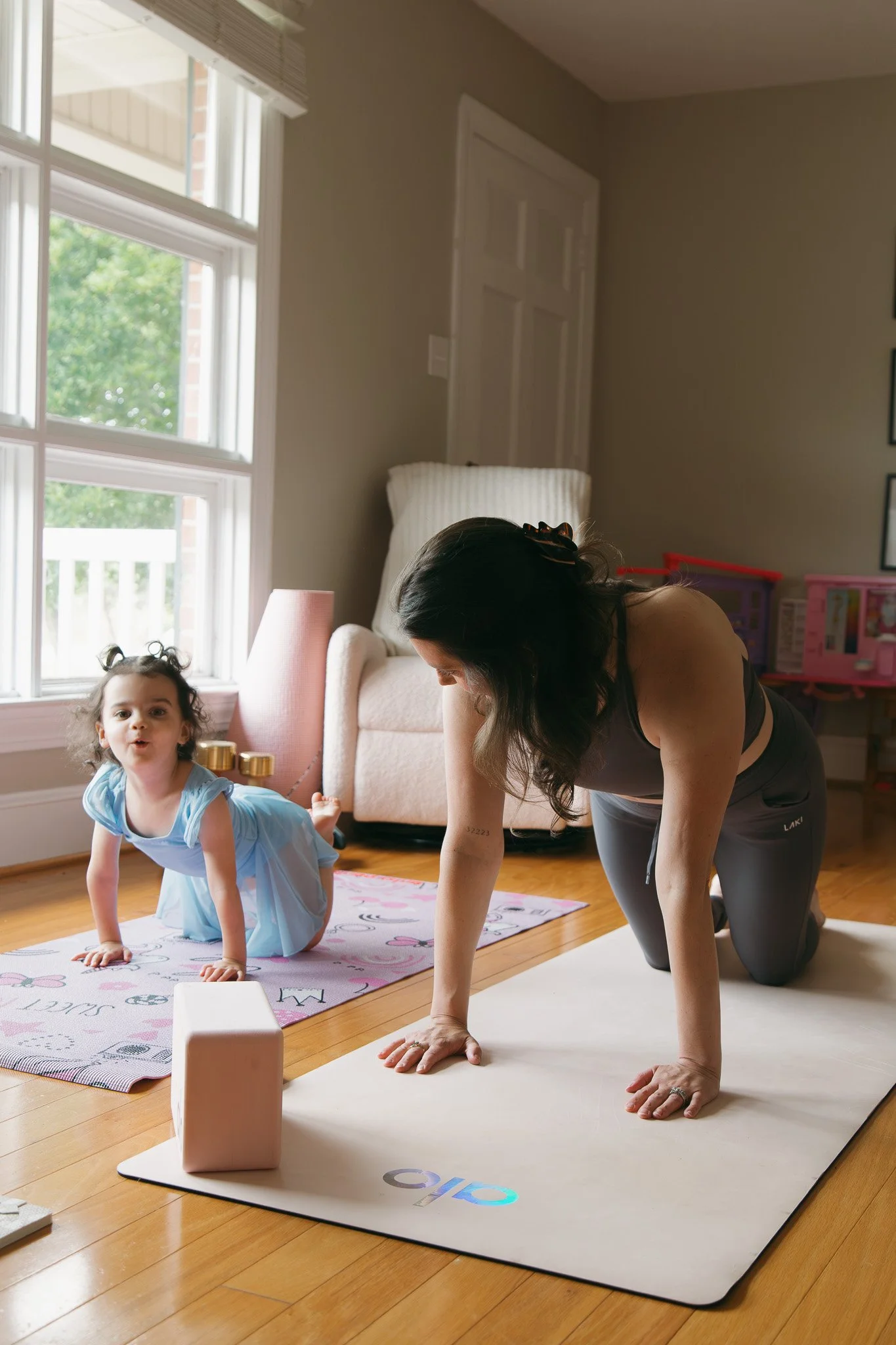 A woman and a young girl practicing yoga in a living room with sunlight coming through the window. The woman is on a yoga mat in a plank position, and the girl, dressed in a blue dress, is on all fours on a nearby rug.