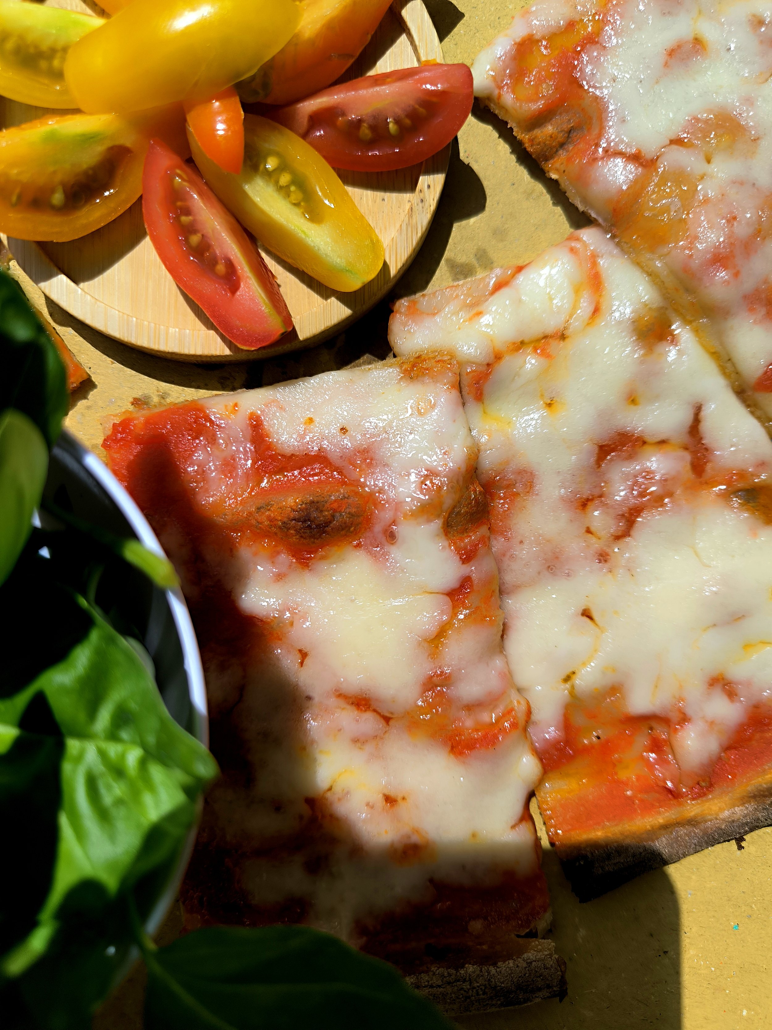 Una fetta di pizza con formaggio e pomodoro, accanto ci sonooni pomodori di diversi colori sopra un tagliere di legno, con alcuni spinaci in primo piano.