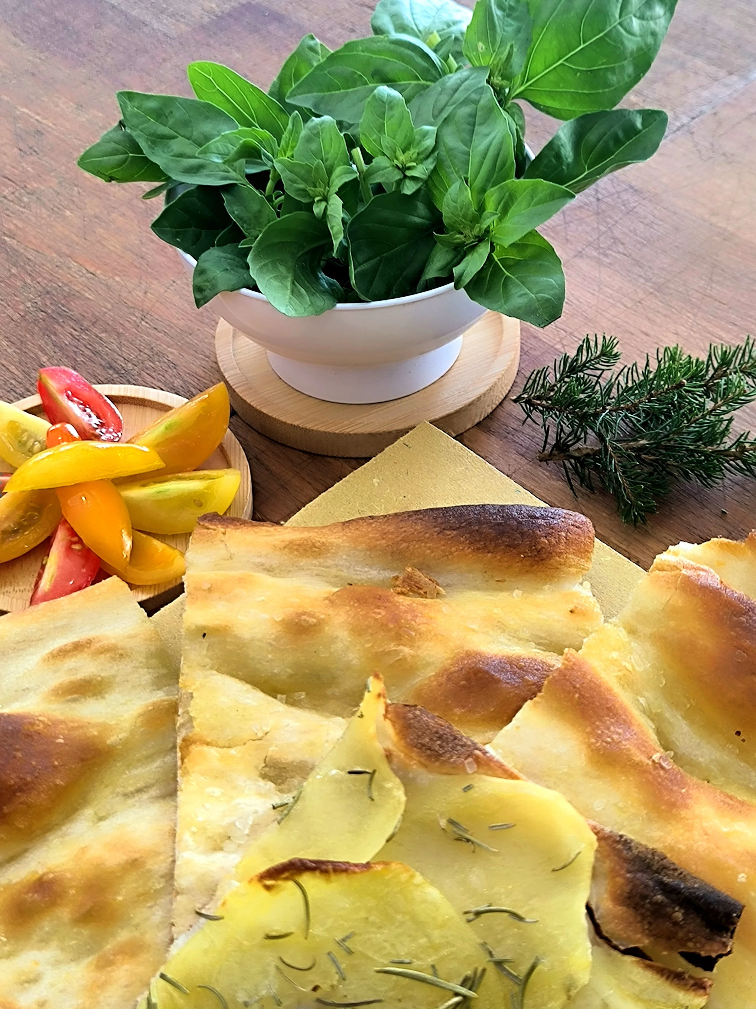 Piatto di pizza con patate e rosmarino, accanto a un piatto di insalata di pomodori, e una pianta di basilico sparse nella scena.