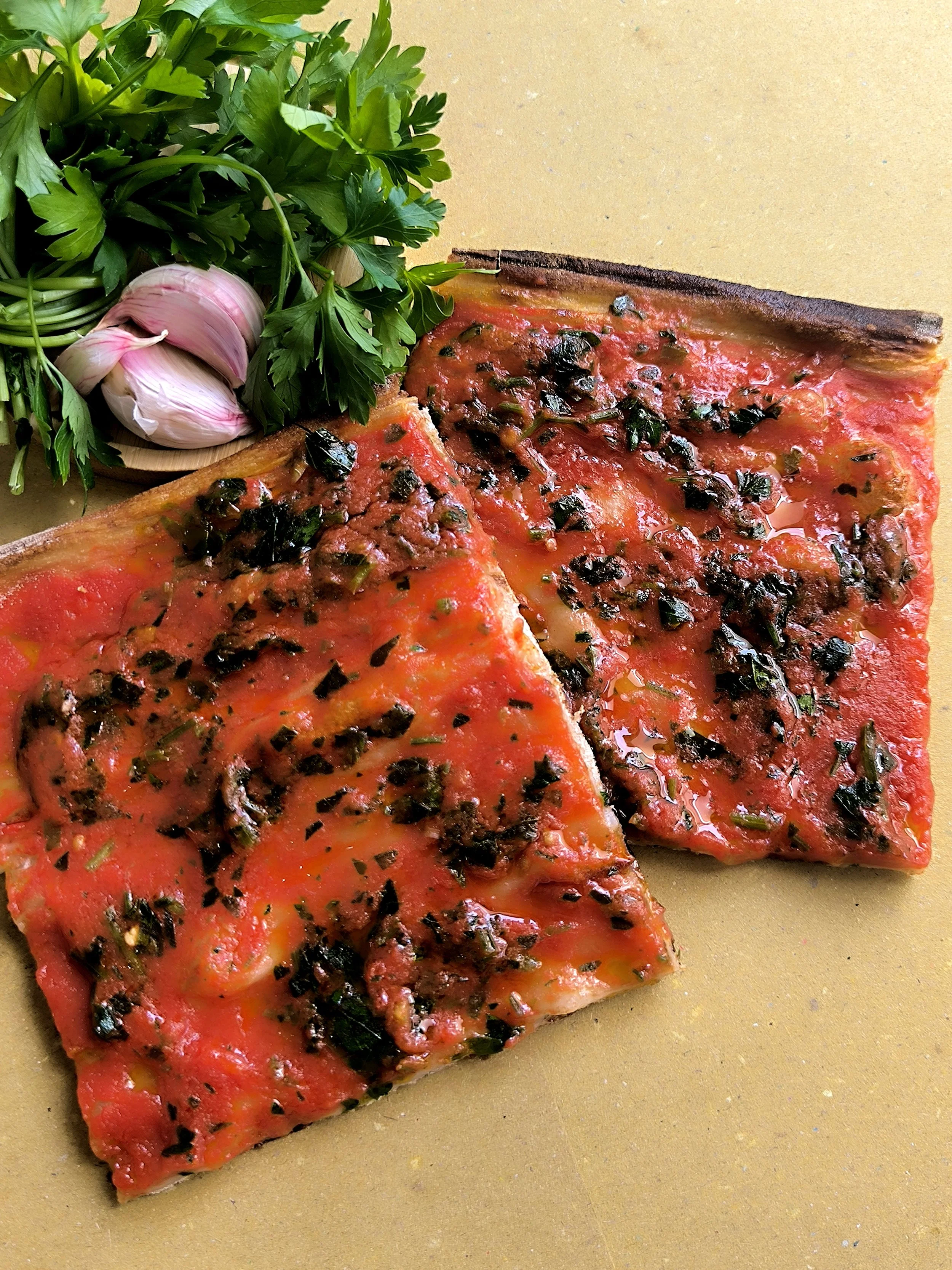 Due fette di pizza con sugo e basilico, accanto a un mazzetto di prezzemolo e due teste d'aglio