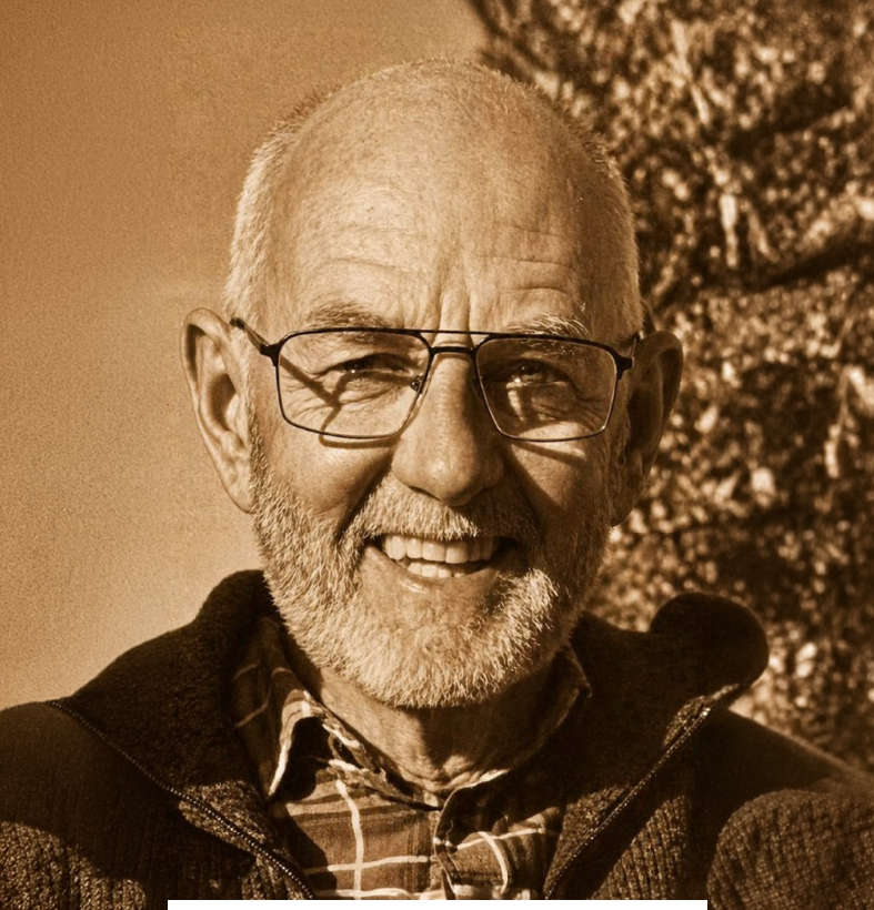 A close-up sepia-toned photo of an elderly man with glasses, a beard, and a smile, wearing a plaid shirt and a jacket, standing outdoors near trees.