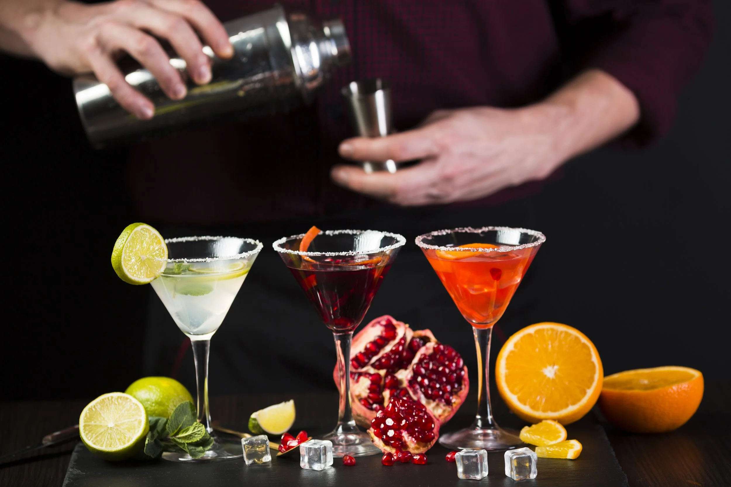 Line of three colorful cocktails in martini glasses with salt rims, surrounded by slices of lemon, lime, and orange, pomegranate seeds, ice cubes, and garnishes on a black background.