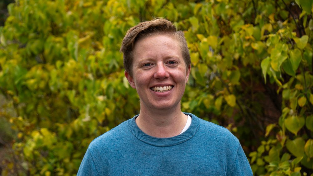 Photo of Court, a white, non-binary person with short blonde hair standing in front of a green, leafy background and wearing a blue sweater. They are smiling at the camera.