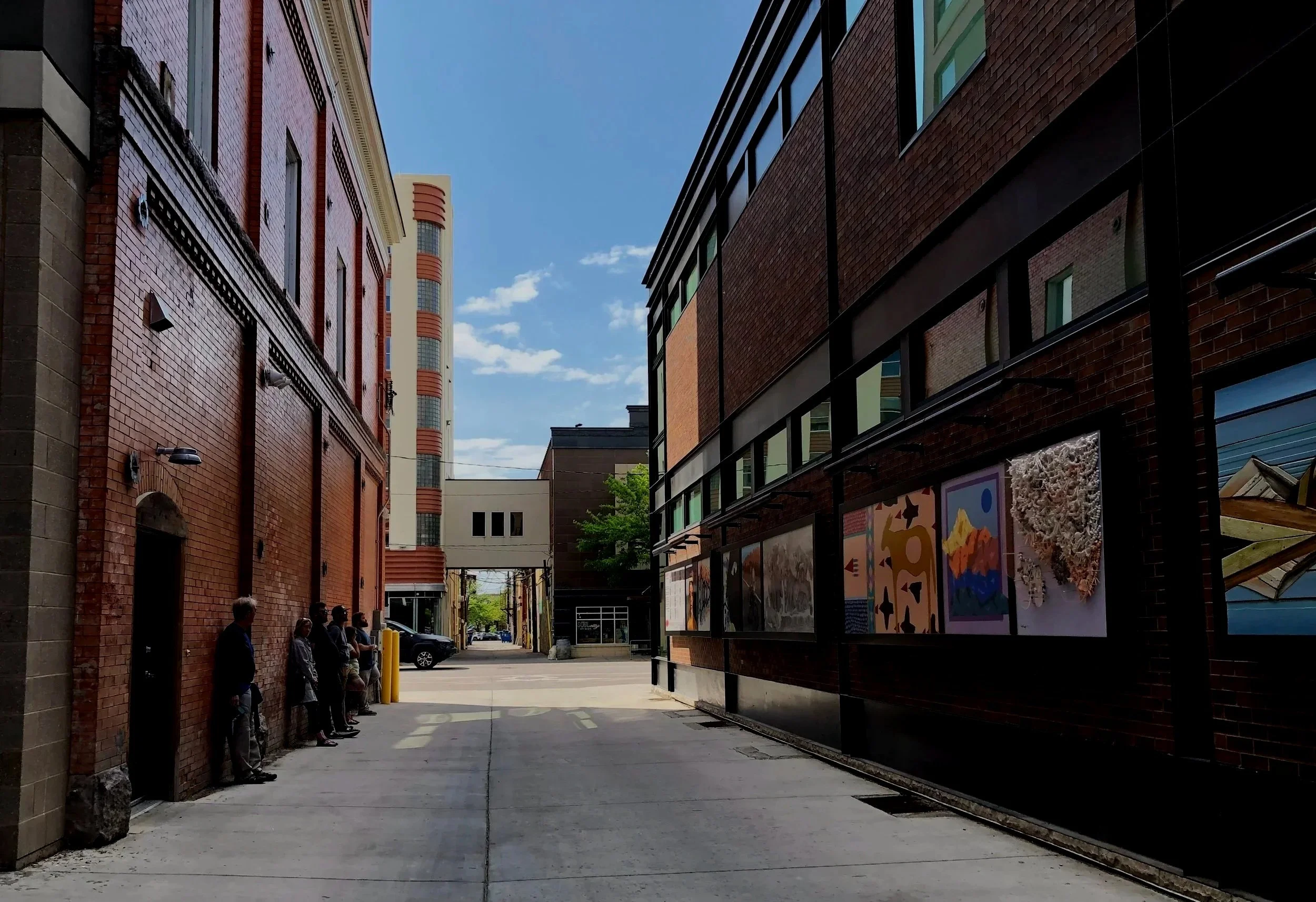Image of an alley way lined with brick walls/buildings. A group of people at Confluence lean against the brick walls on the left side of the photo and gaze at an outdoor art gallery of photos and painting on the right side of the alleyway.