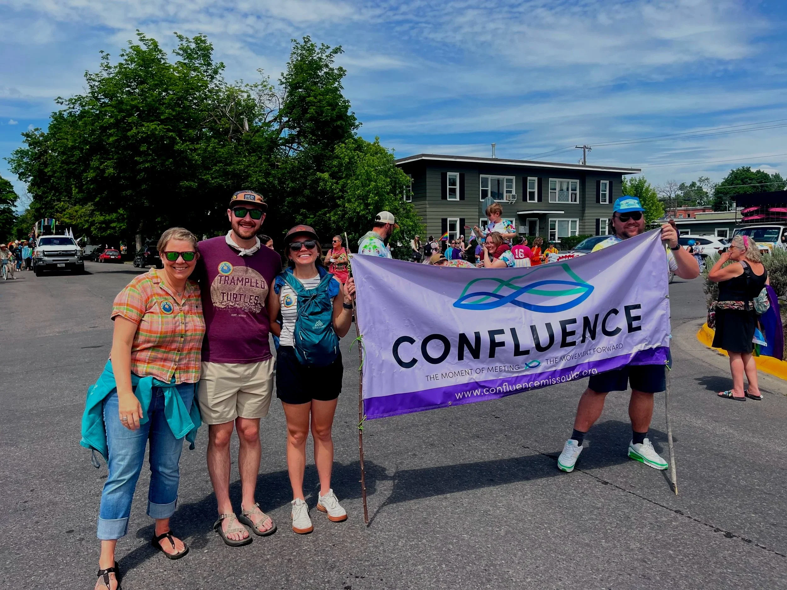 Three people stand close together. The one on the right holds a purple "Confluence" Banner, while another person holds the other side of the banner. These individuals are at a pride parade.